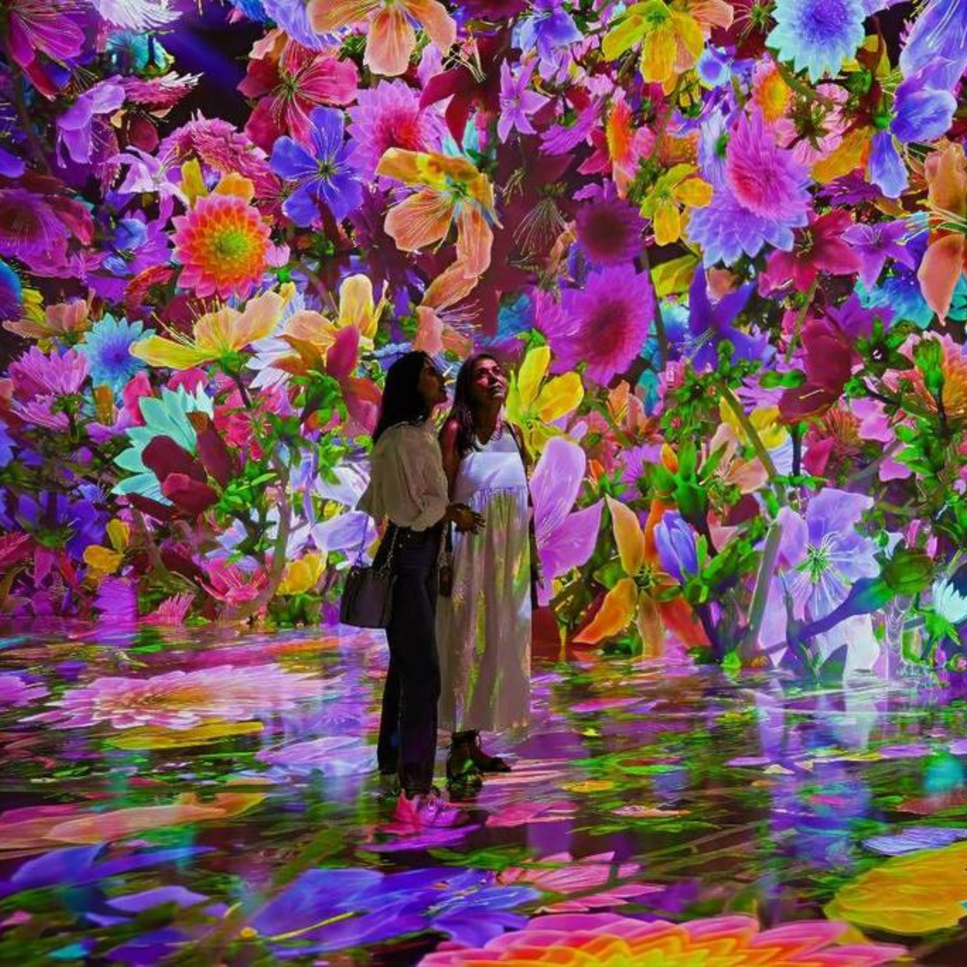 Two people stand inside an immersive digital art installation where vibrant multicolored flowers bloom across walls and floor, creating a surreal, kaleidoscopic environment