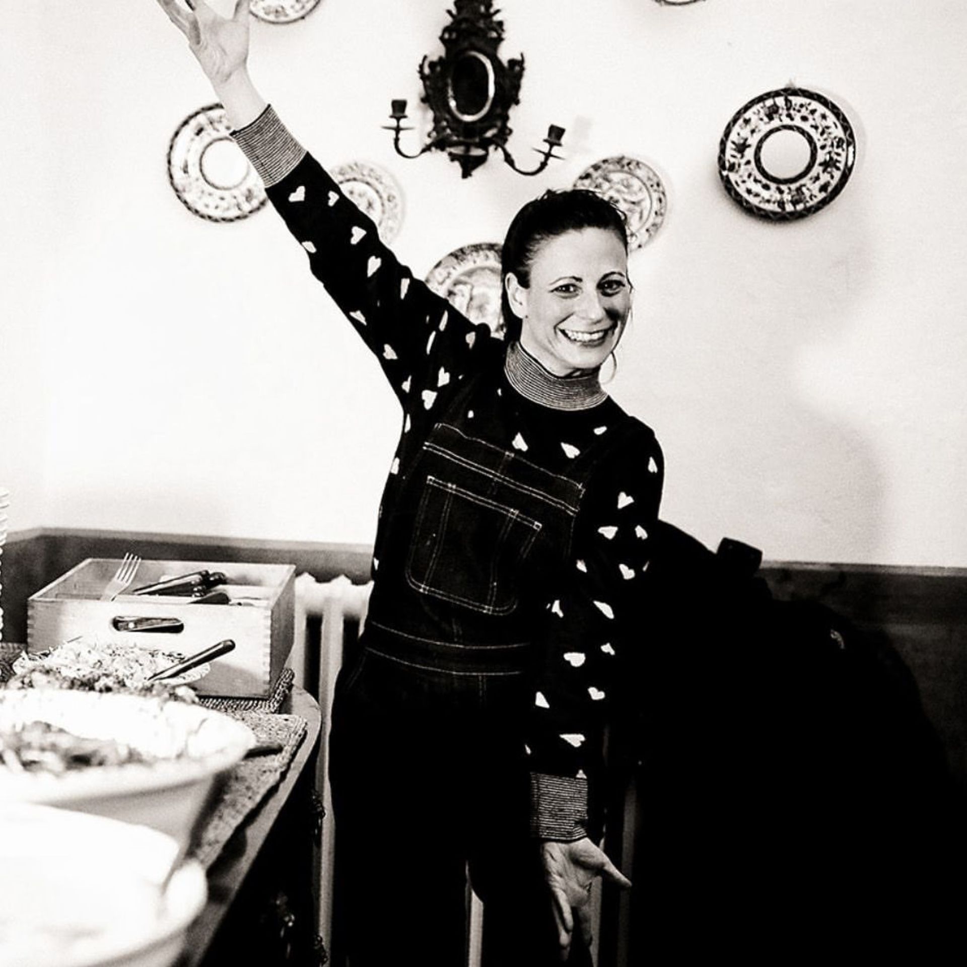 Person in heart-patterned top and overalls smiling indoors with decorative plates and dining table in background