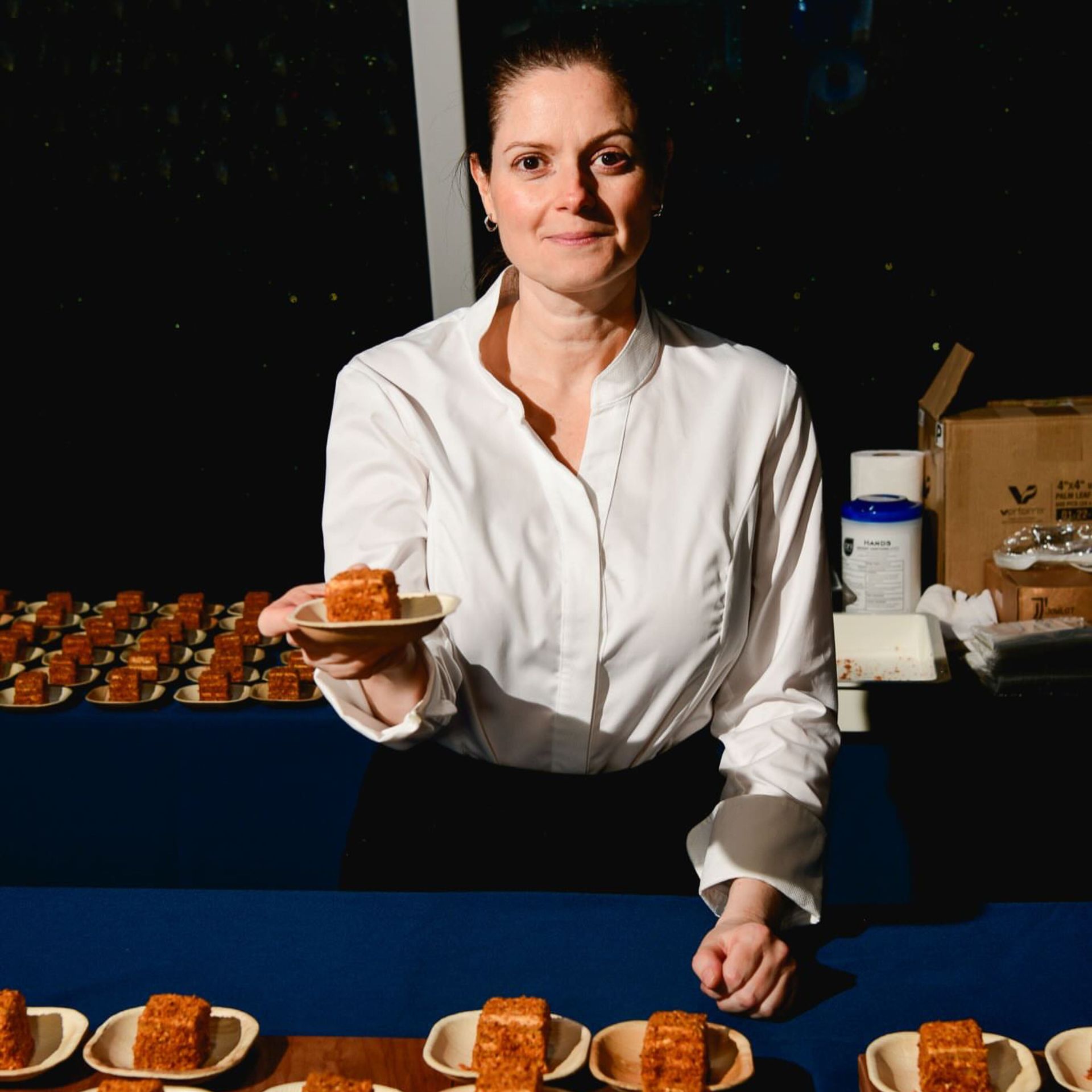 Chef in white coat presenting plated dessert squares at table with multiple servings arranged in rows