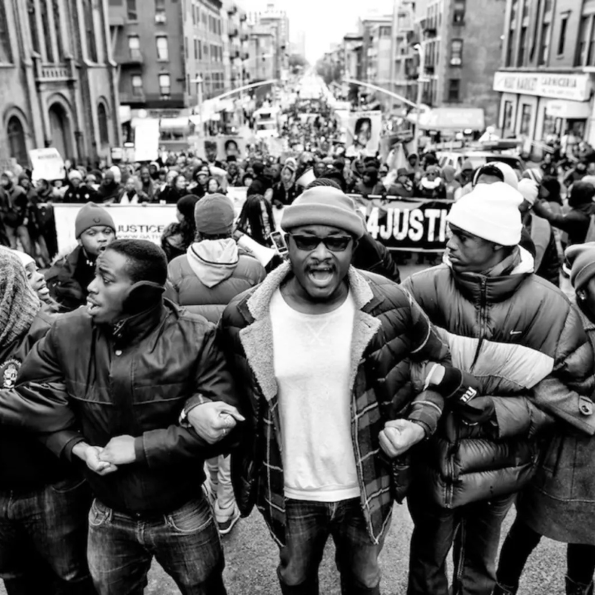 Black-and-white photo of a crowded protest march, with people linking arms and a banner reading Justice visible in the background