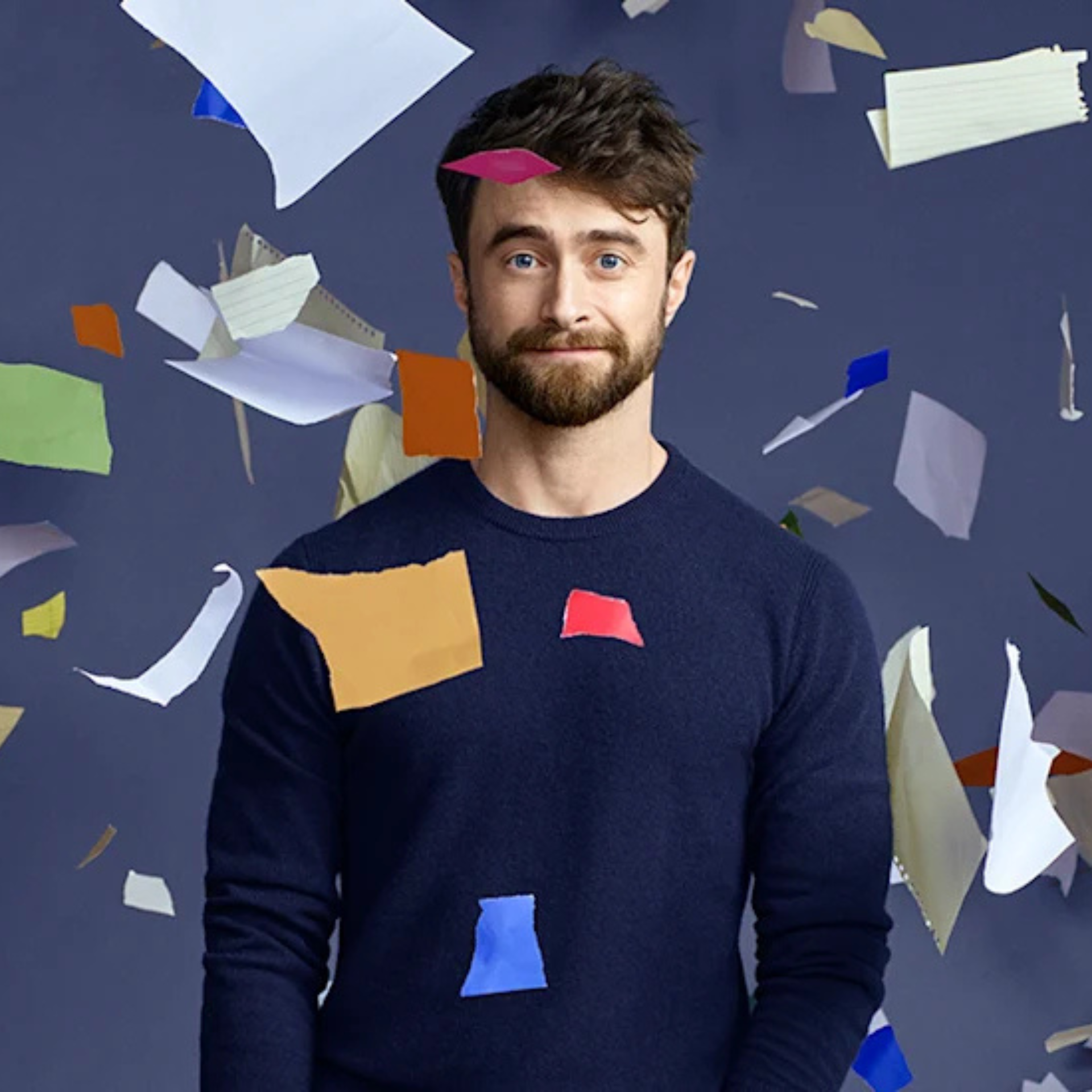 A person in a dark blue sweater stands against a black background as colorful sheets of paper float mid-air around them, creating a dynamic, chaotic atmosphere