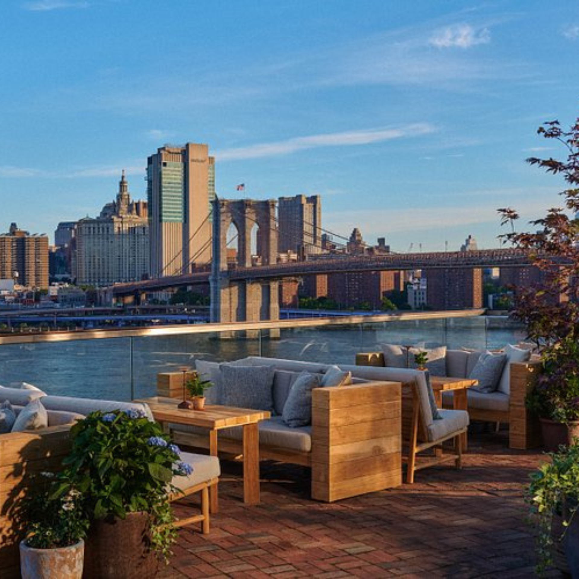 Rooftop terrace with wooden furniture, potted plants, and a view of the Brooklyn Bridge and East River skyline