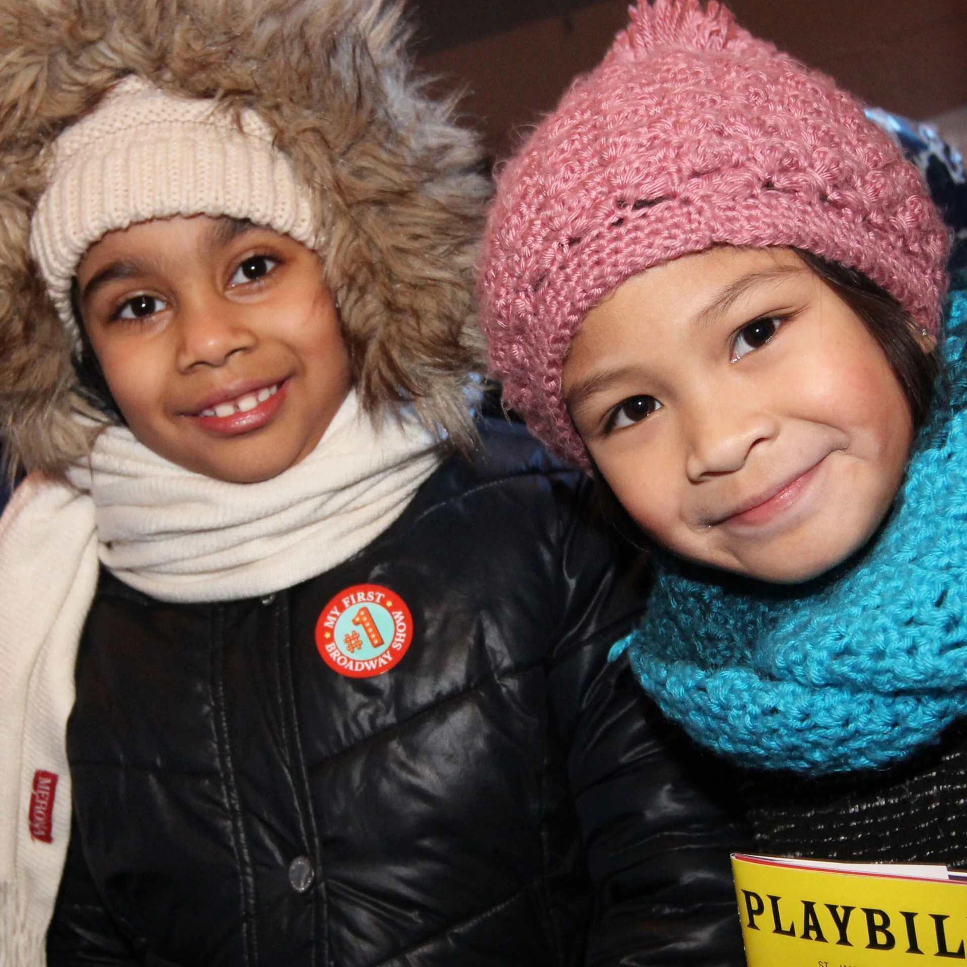 Two children in winter hats and scarves smiling after their first Broadway show, holding a Playbill and wearing a badge