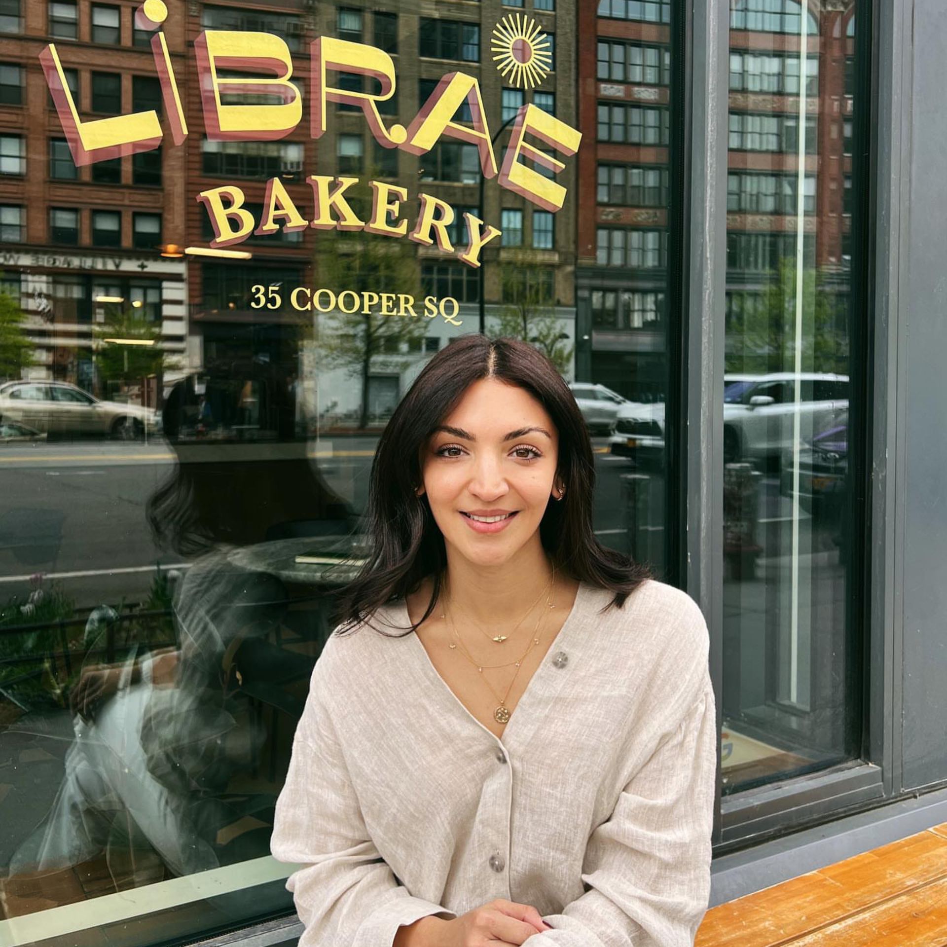 Person smiling on bench outside Librae Bakery with glass window signage and street reflections in background