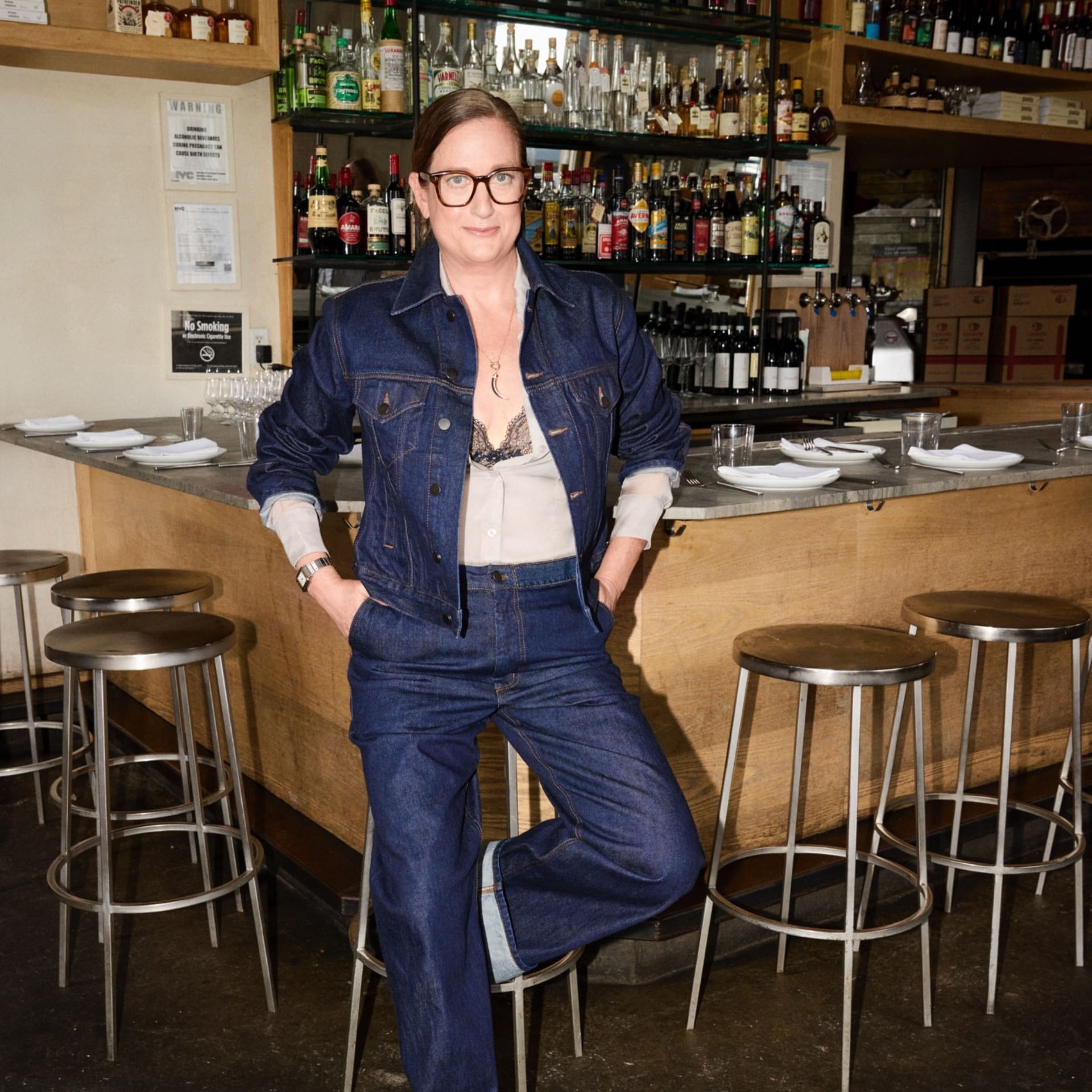 Person in denim jacket and sheer blouse standing at bar counter with bottles and stools in background