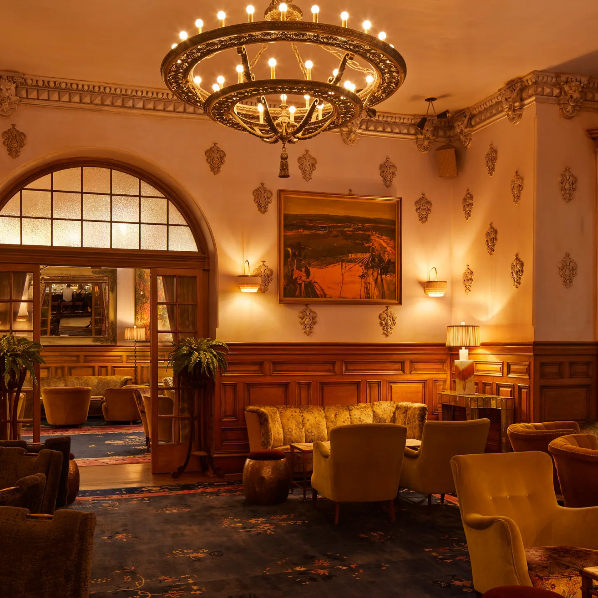 Warmly lit lounge with chandelier, patterned wallpaper, vintage seating, and arched doorway leading to another room