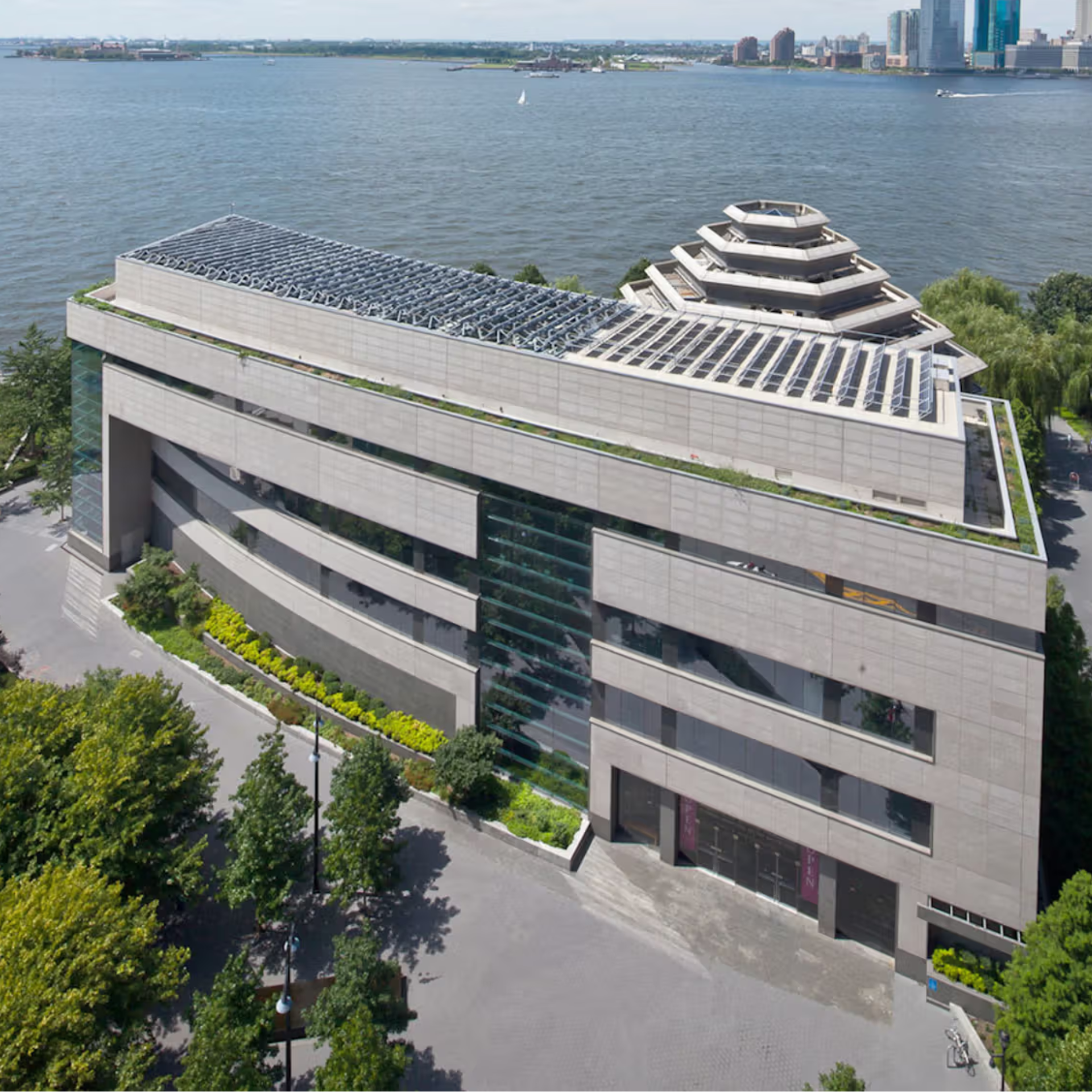 Exterior view of the Museum of Jewish Heritage in Lower Manhattan, featuring its six-sided tiered roof with solar panels, landscaped greenery, and Hudson River backdrop