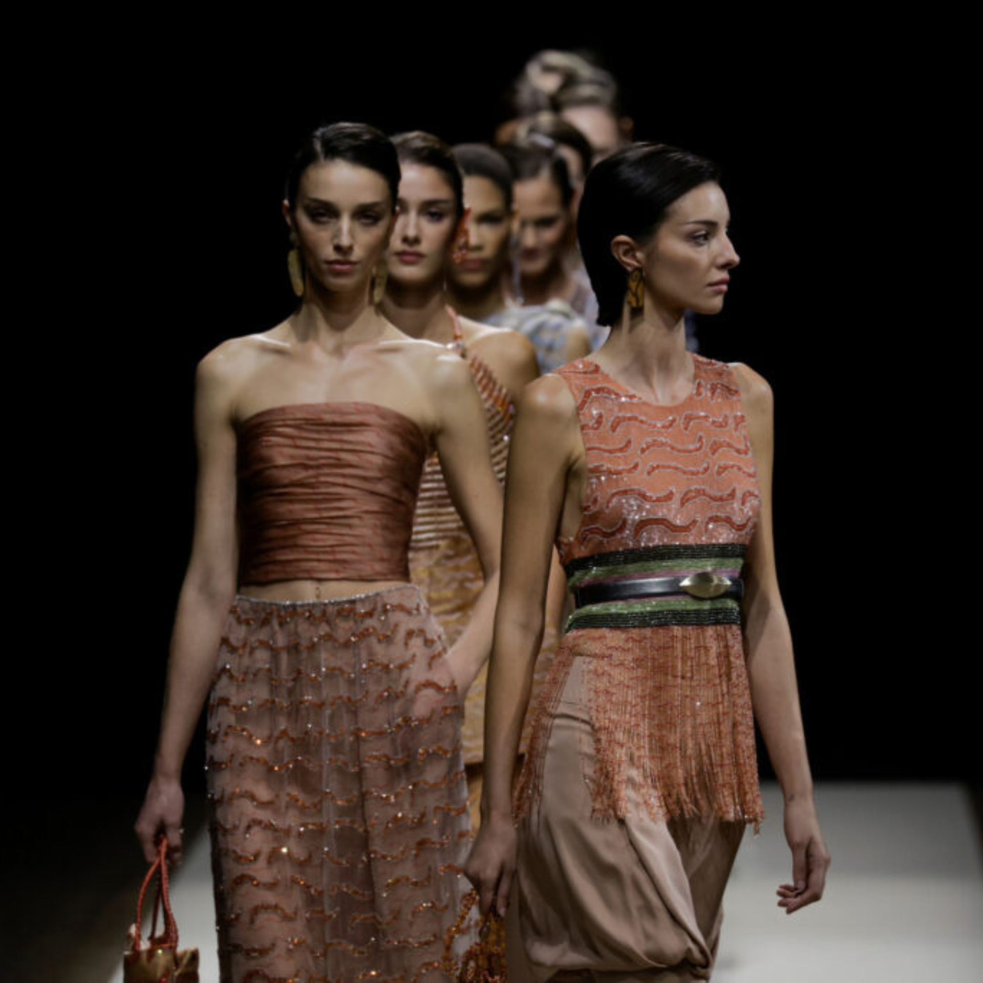 Runway models in earthy-toned, textured outfits with metallic accents at a high-fashion show