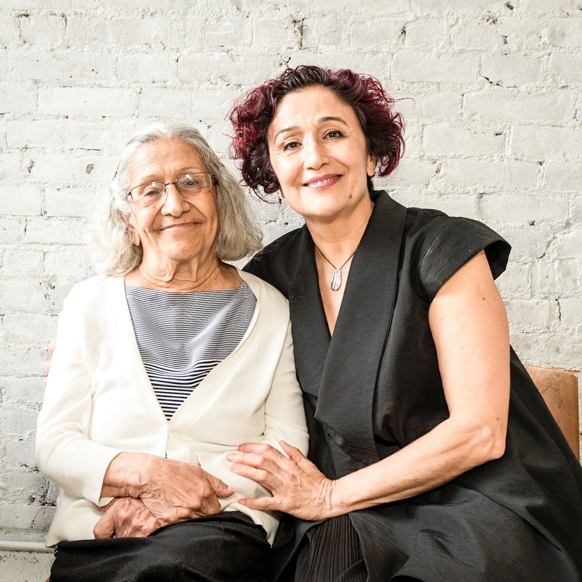 Two people sitting together, smiling in front of white brick wall, one with gray hair and glasses, the other with short curly hair and red highlights