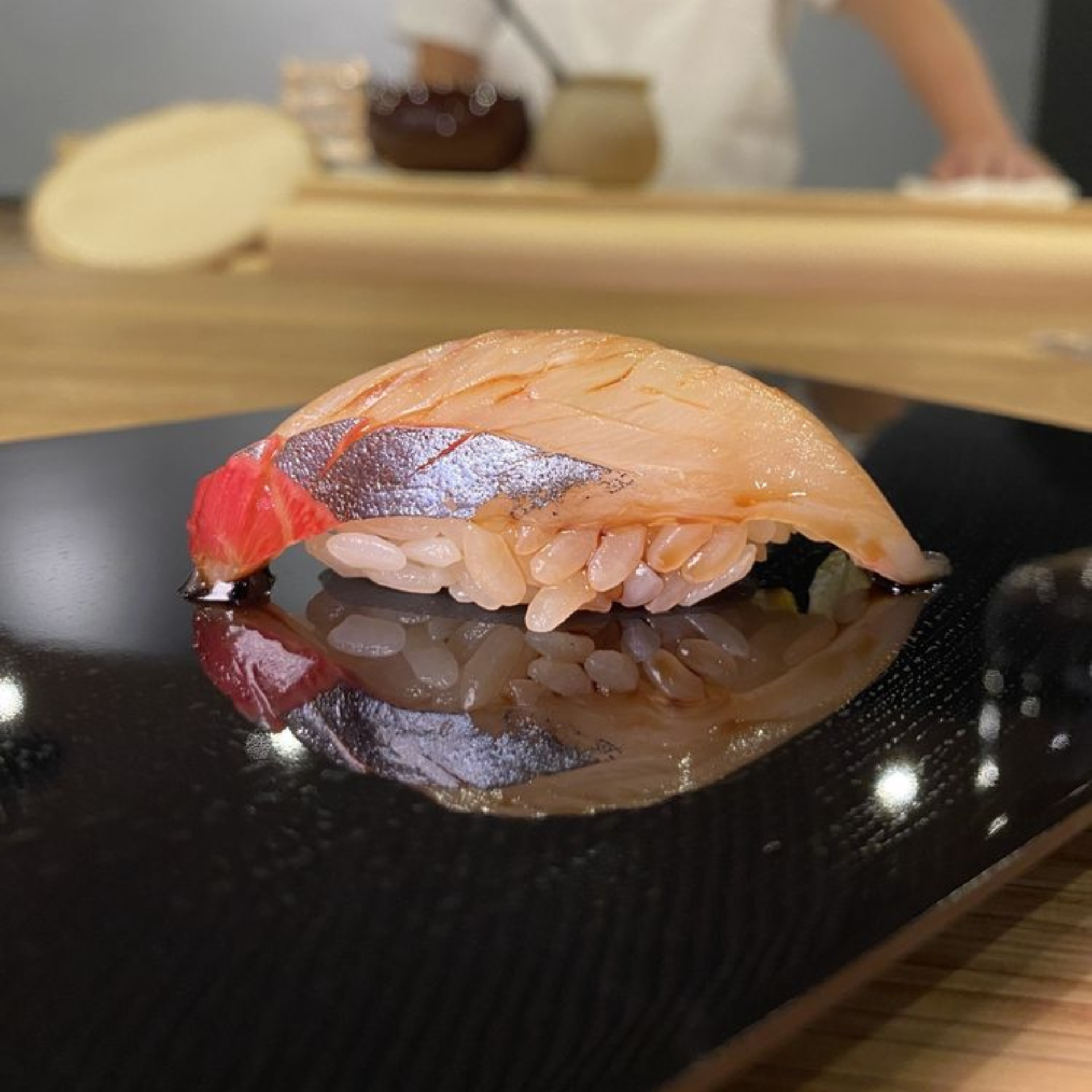 Piece of sushi with shiny silver fish topping on rice, served on a black plate at a sushi bar
