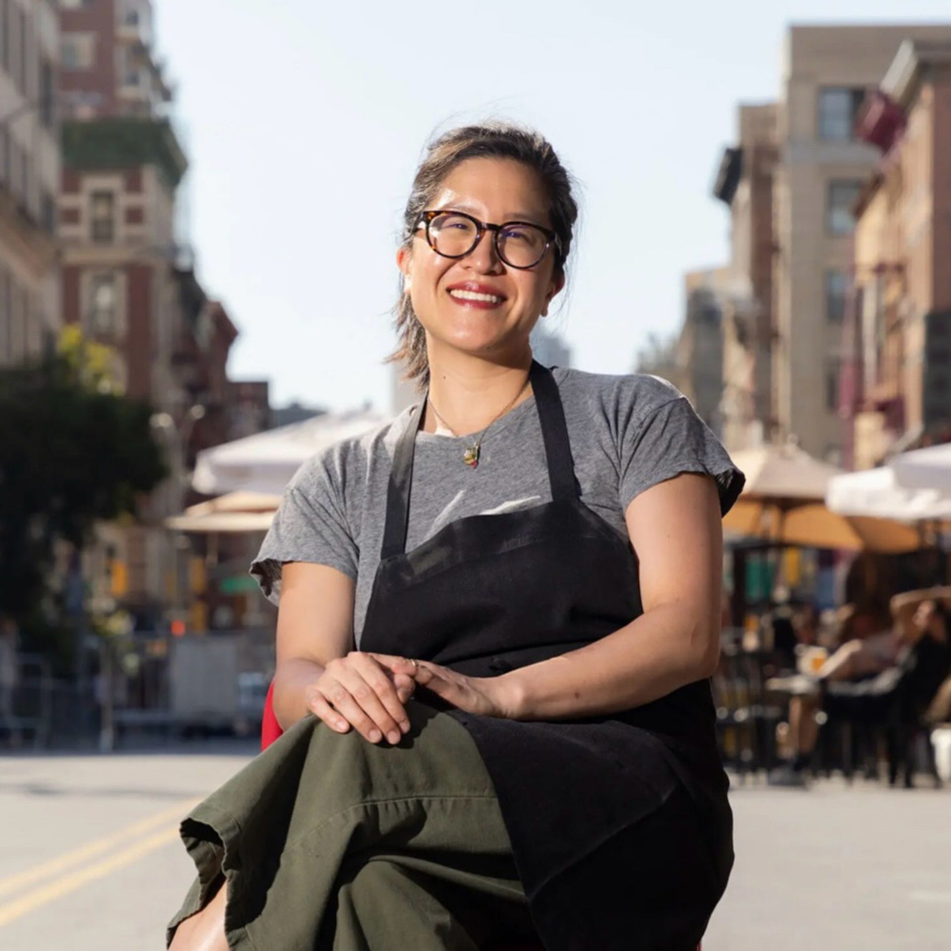 Person in gray t-shirt, black apron, and green pants smiling while seated outdoors on city street with buildings and umbrellas in background