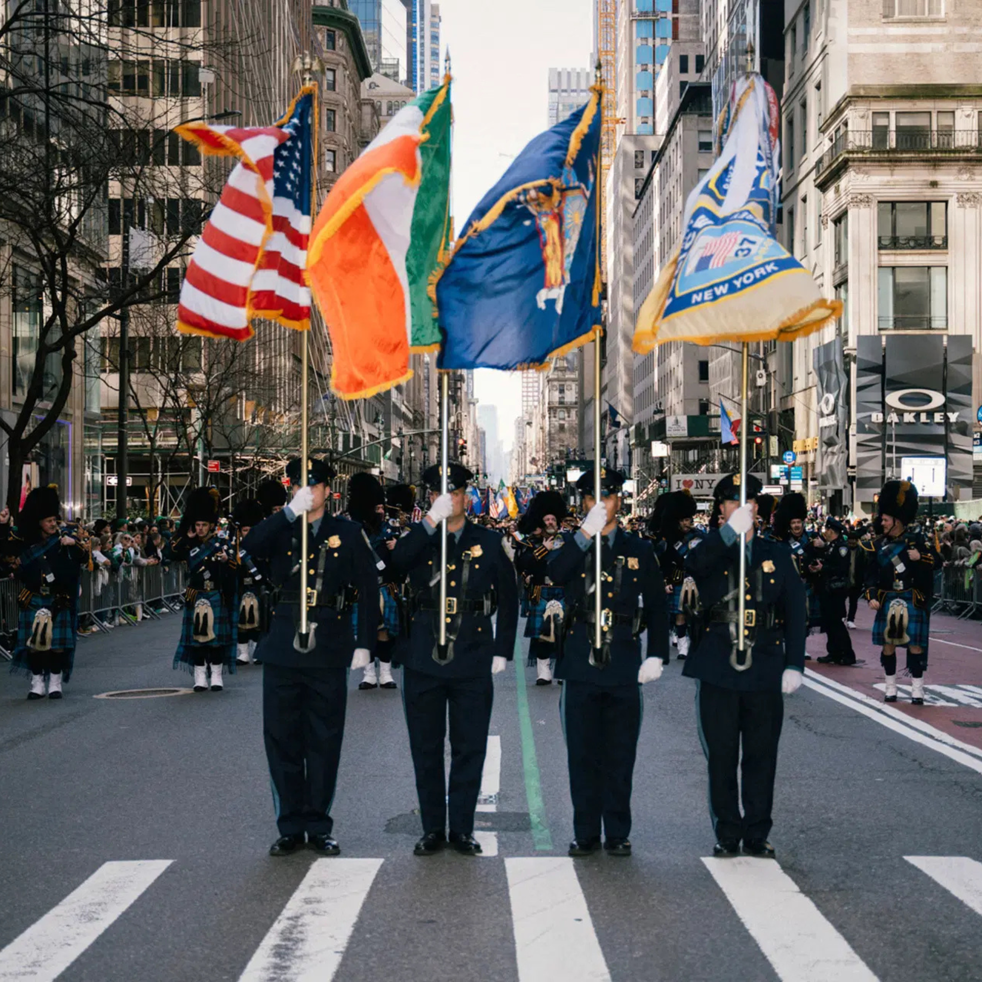 Uniformed officers carry American, Irish, New York, and blue flags at the front of a city parade, with a marching band in kilts and spectators lining the street