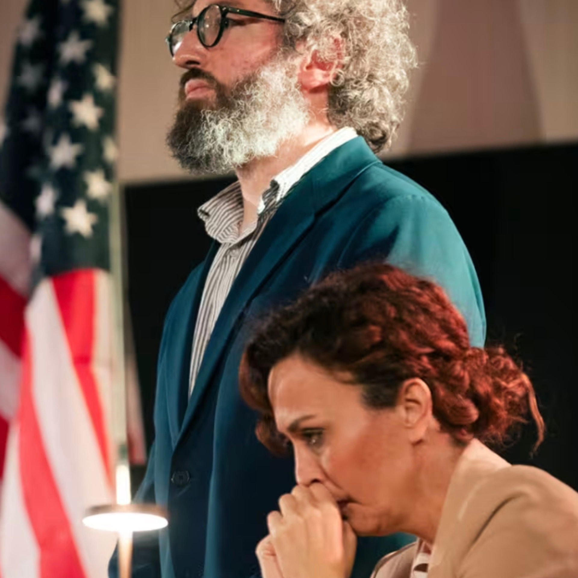 Two individuals indoors with an American flag in the background; one seated, appearing contemplative, and another standing in a suit with a serious expression