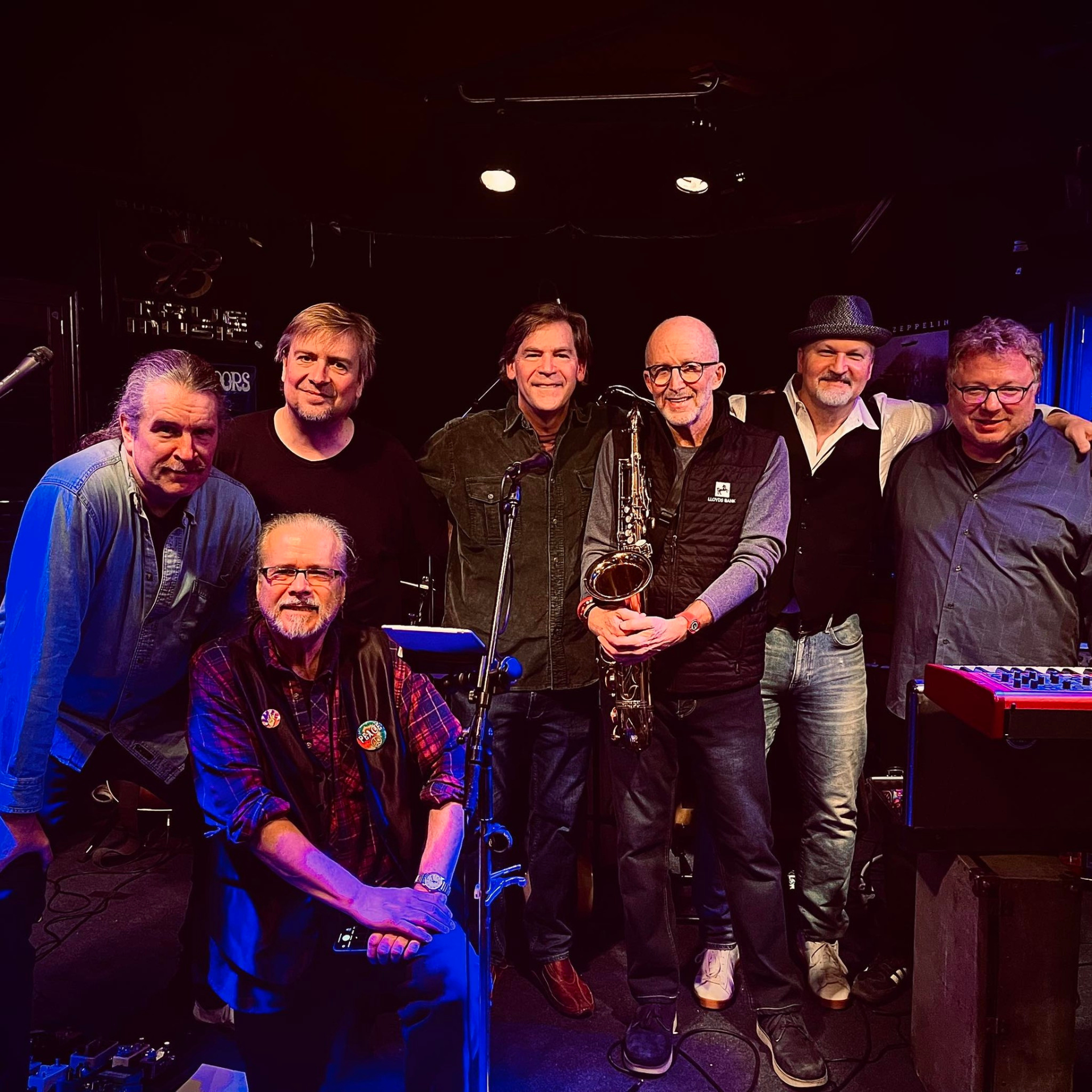 Seven musicians posing together on stage with instruments after a live performance