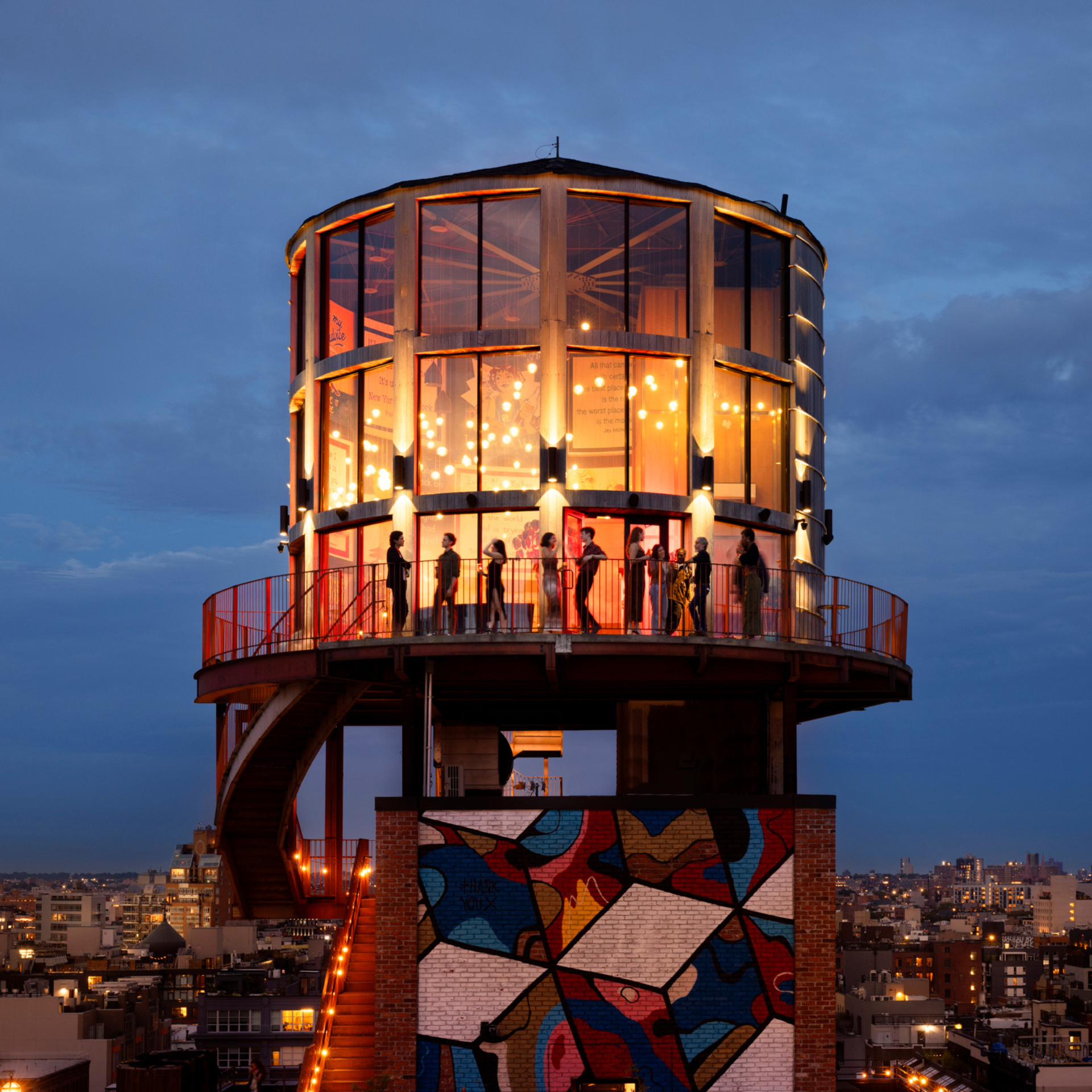 Cylindrical glass room glowing at dusk above a mural-covered building with city skyline views