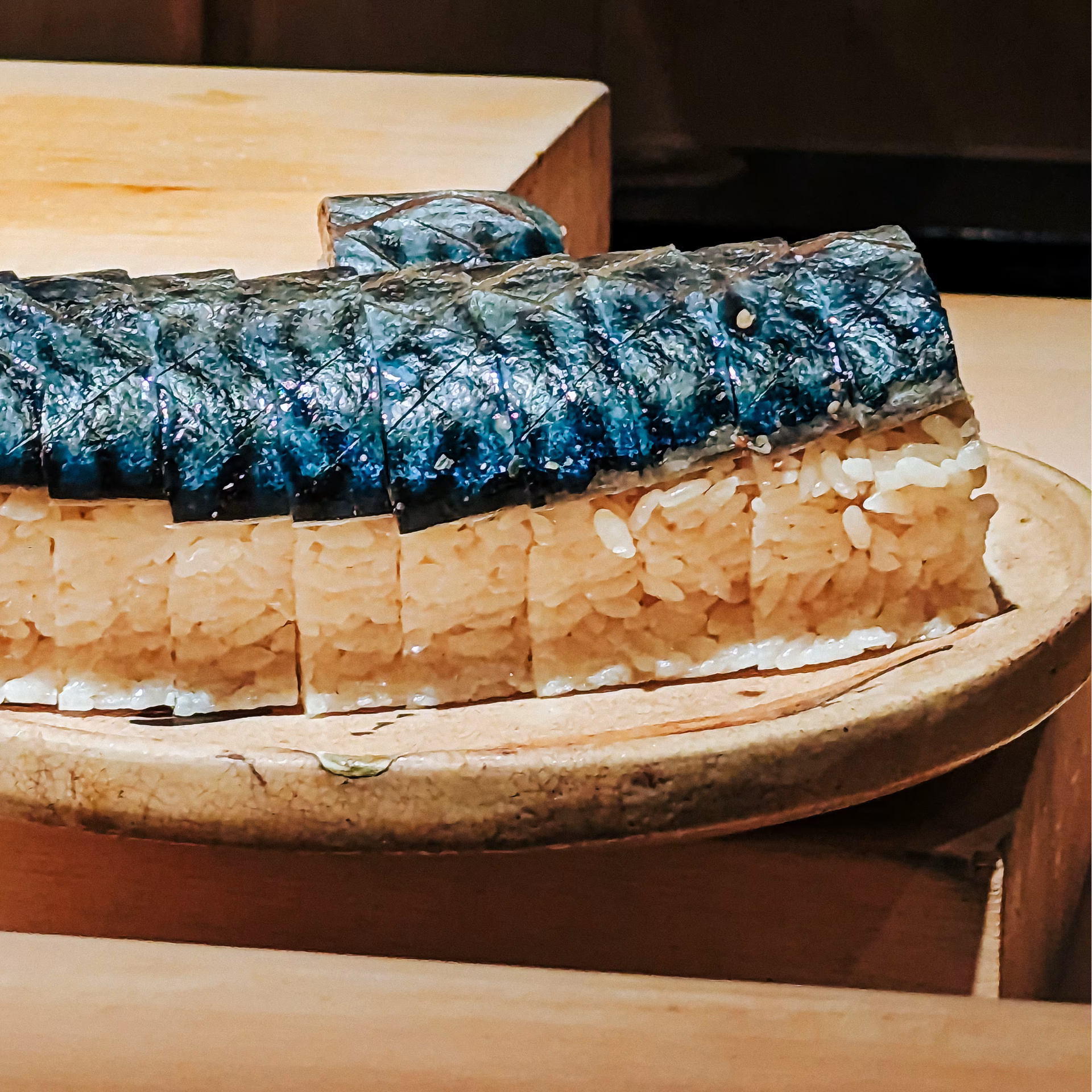 Rectangular pressed sushi with shiny mackerel topping, sliced and served on a wooden board