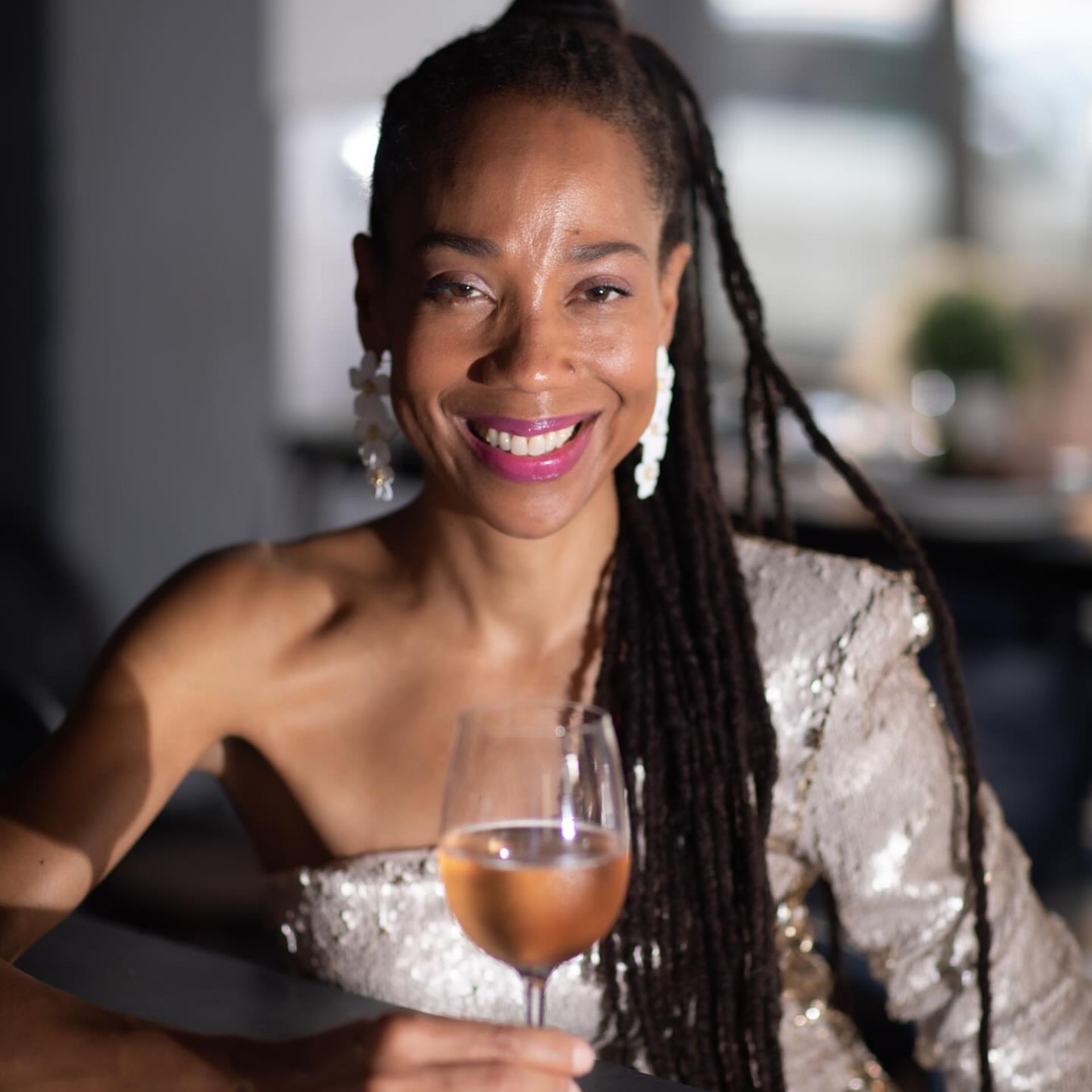 Person in silver one-shoulder outfit with braided hair and floral earrings, smiling while holding a glass of rosé indoors