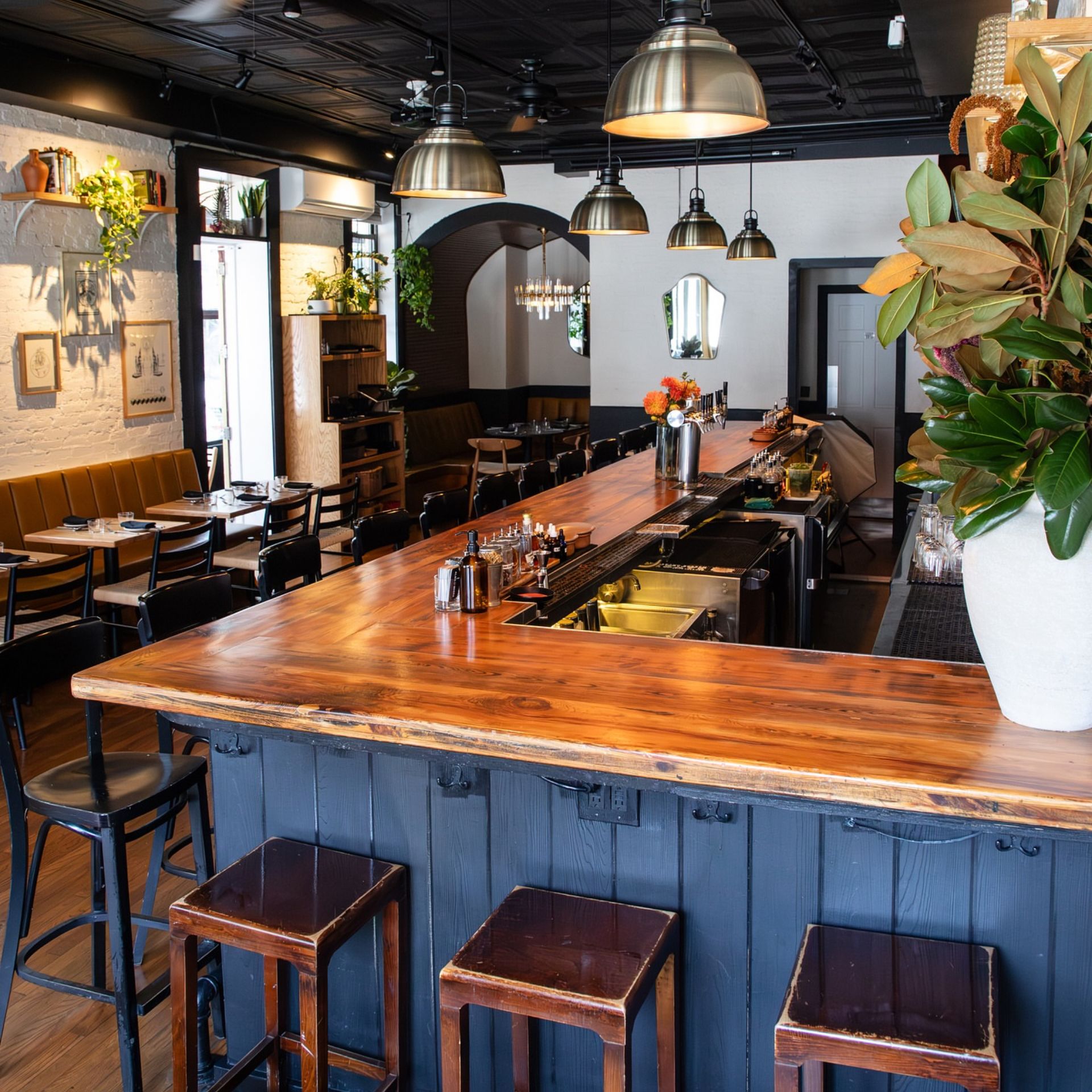 Modern restaurant interior with L-shaped wooden bar, stools, pendant lights, and cozy seating with framed artwork and plants