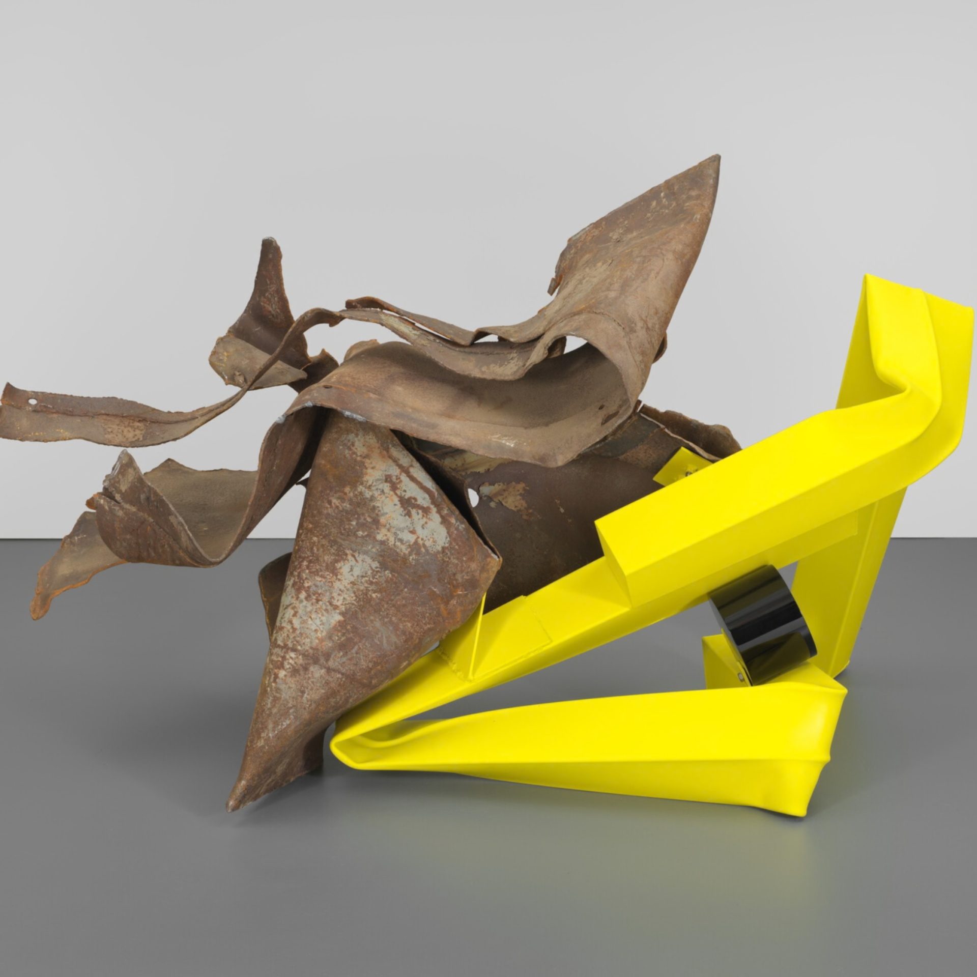 Abstract sculpture with rusted twisted metal atop bright yellow geometric base against gray wall