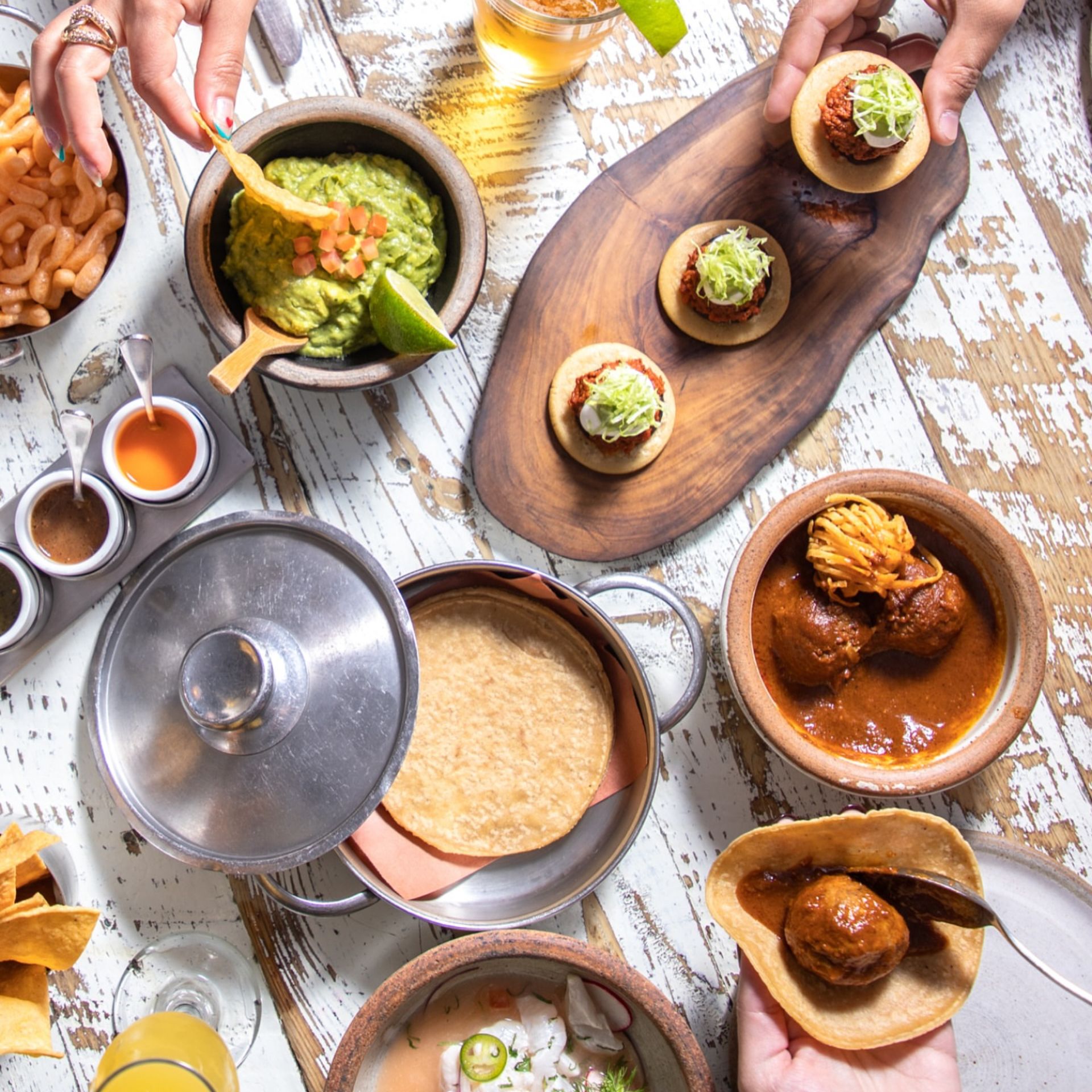 Rustic table spread with Mexican dishes including guacamole with chips, tostadas, meatballs in red sauce, ceviche, tortillas, hot sauces, and drinks