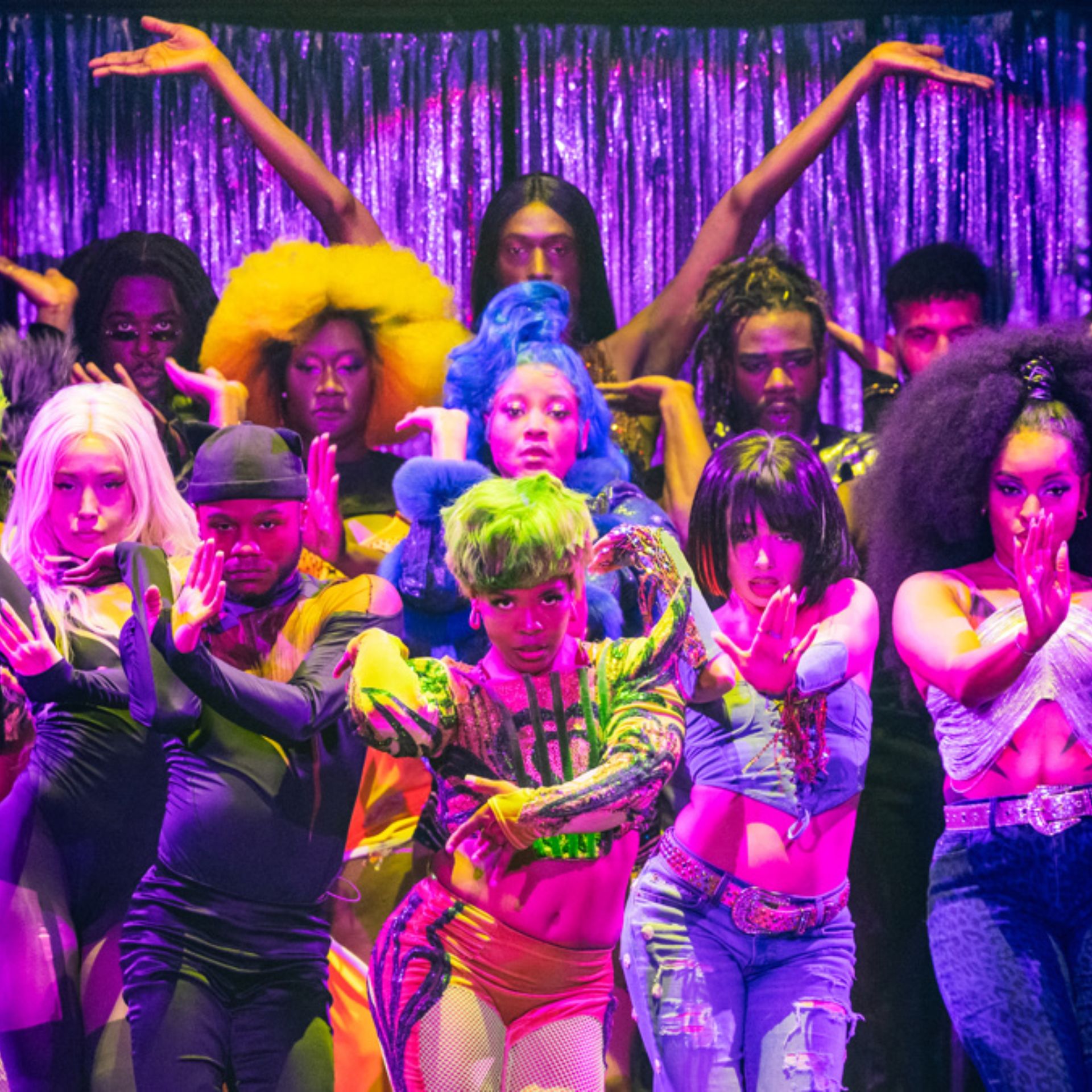 Performers in colorful wigs and costumes strike expressive poses on stage against a shimmering purple curtain