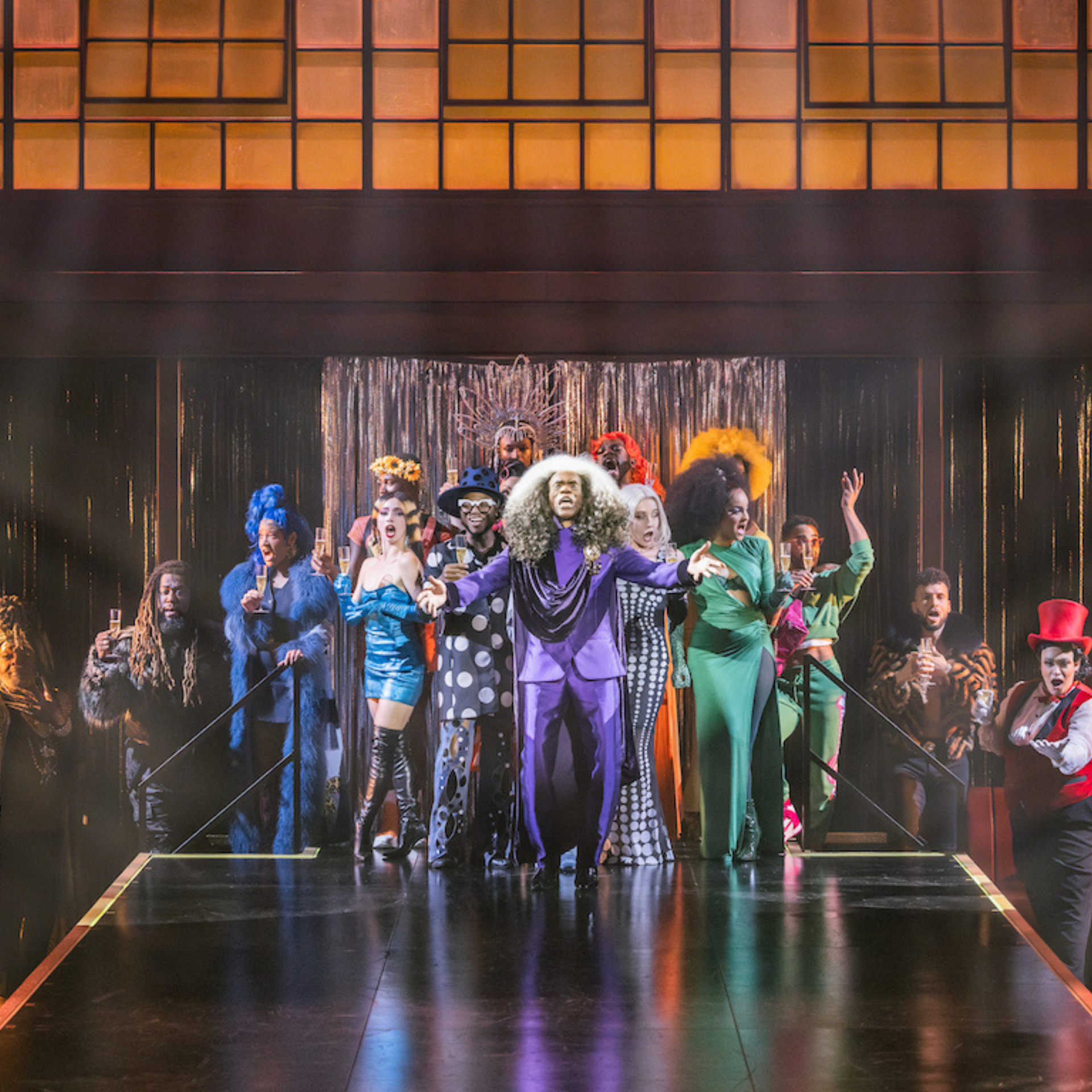 Performers in vibrant costumes on stage, central figure in purple suit with silver wig raising arms, others holding glasses in celebration
