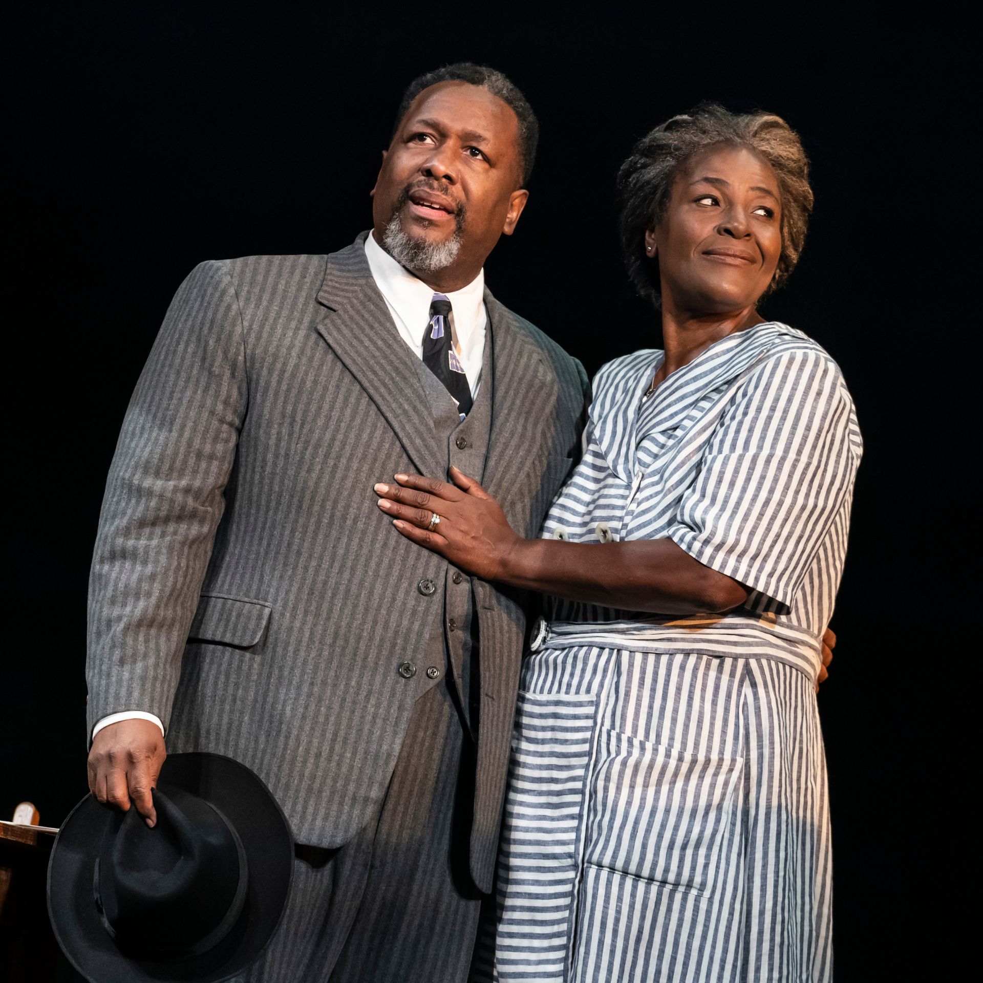 Two actors on stage, man in pinstriped suit holding a hat, woman in striped dress with hand on his chest, both gazing upward against a dark backdrop