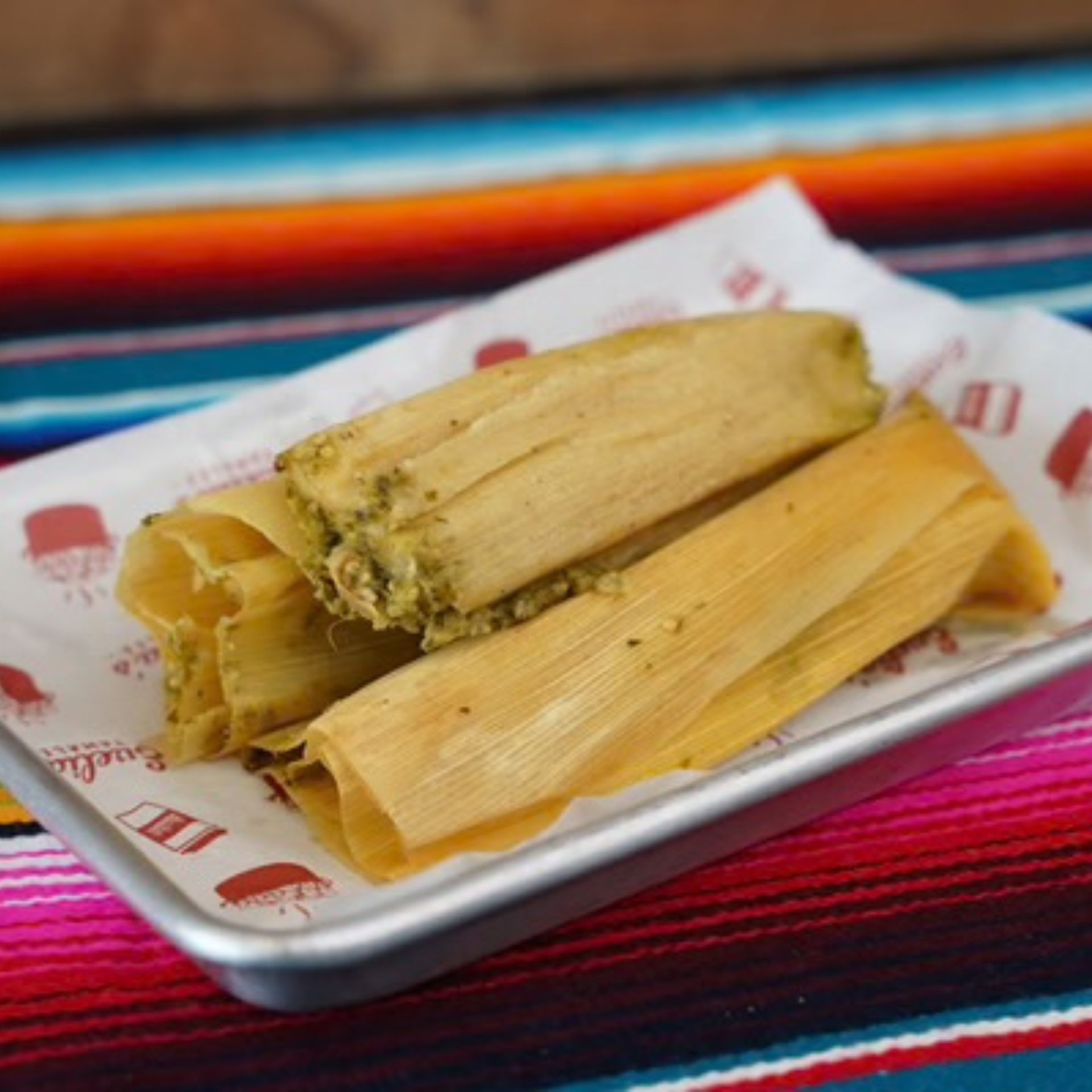 Plate with three tamales wrapped in corn husks on tray with branded paper, set atop colorful striped textile