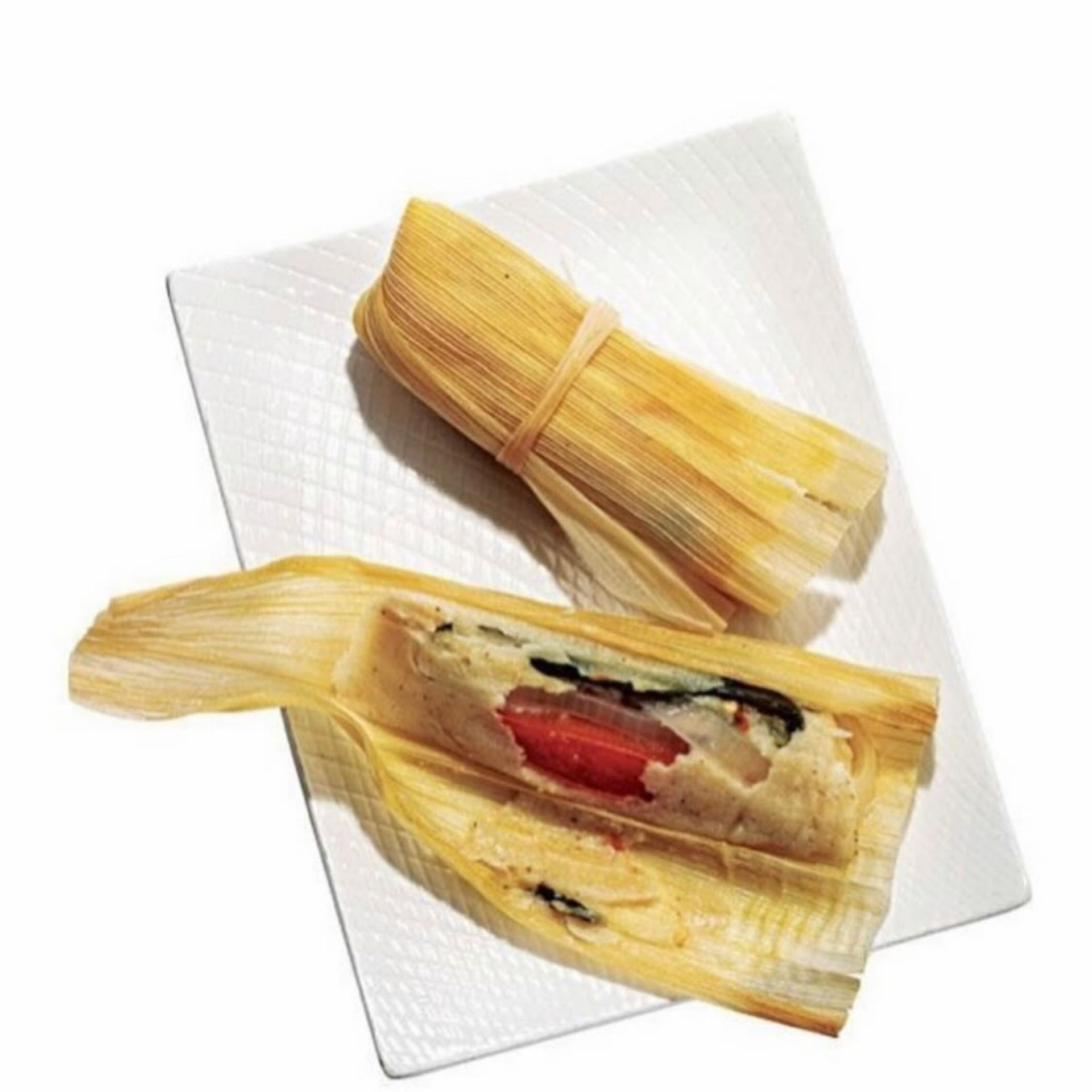 Two tamales on white square plate, one wrapped in husk and one opened to show filling with chili pepper and masa