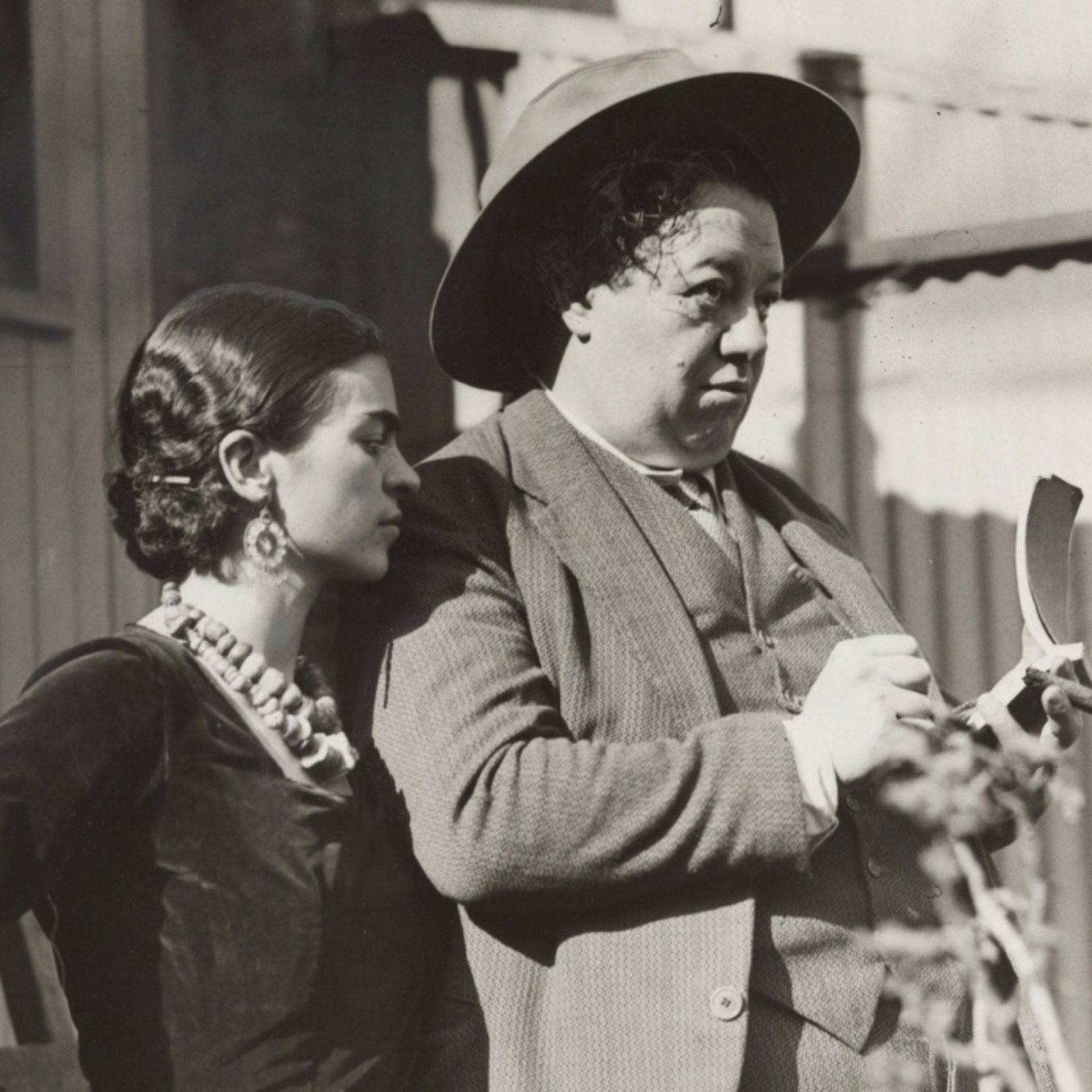 Black-and-white photo of two people standing close together, one in a dress with jewelry, the other in a suit and hat holding a ceramic piece