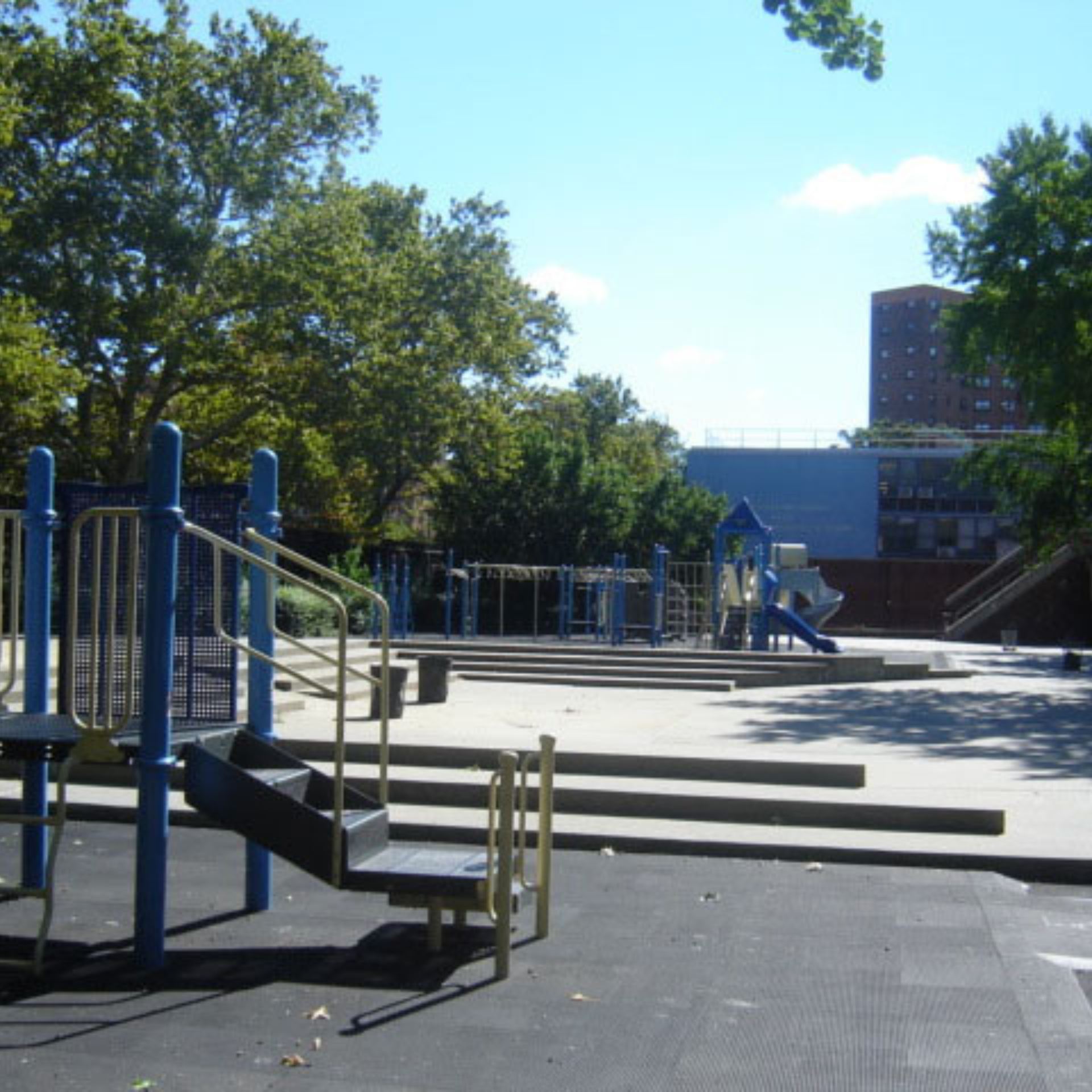Urban playground with blue and beige slides, climbing structures, and safety matting surrounded by trees