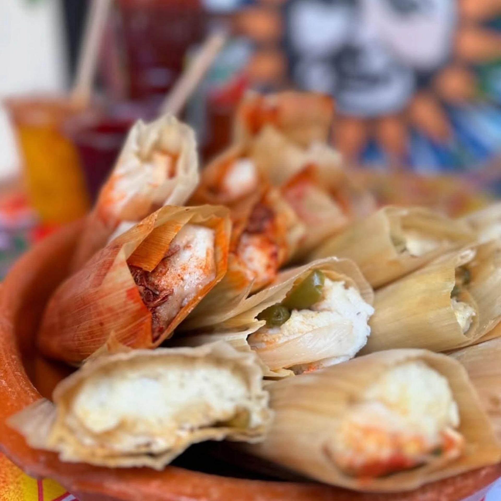 Clay dish with tamales wrapped in corn husks, filled with chili sauce, peppers, and cheese, set against colorful drinks and mural background