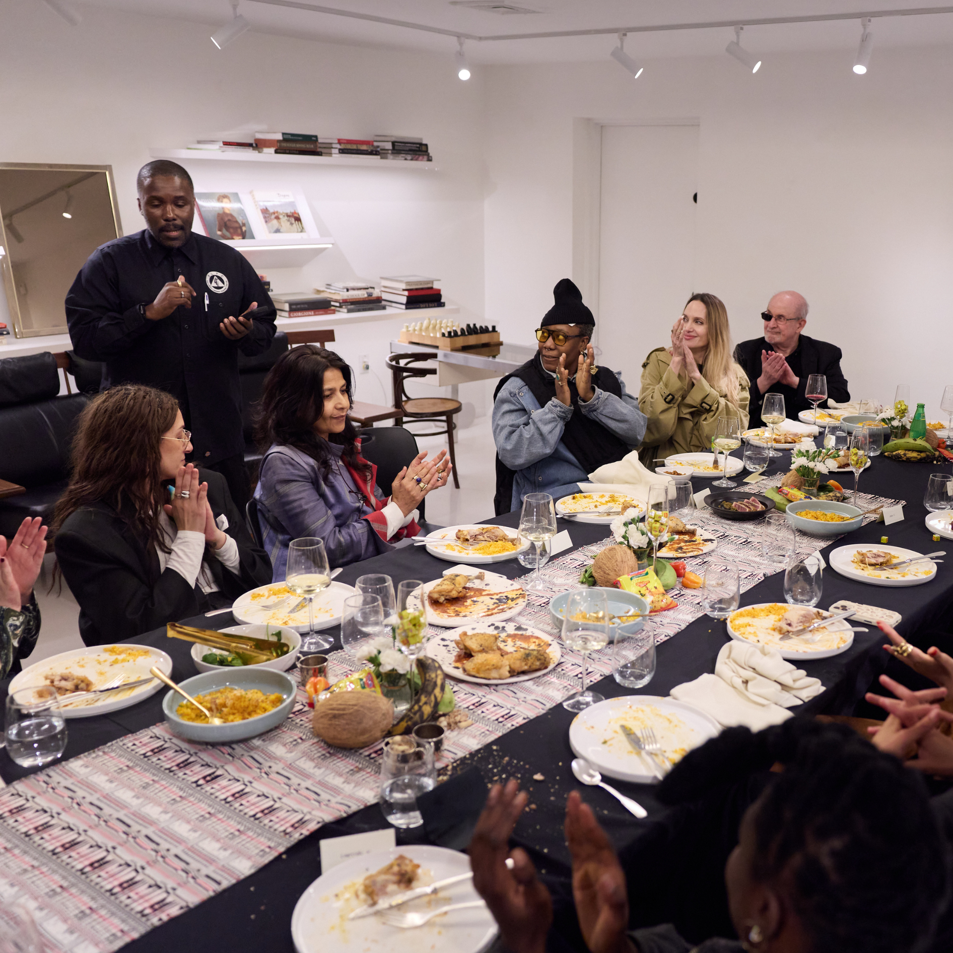 Group of people seated at a long dining table with food, drinks, and decorations in a modern room