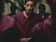 Man in maroon scrubs stands in a dim corridor, hands clasped, surrounded by others in similar uniforms