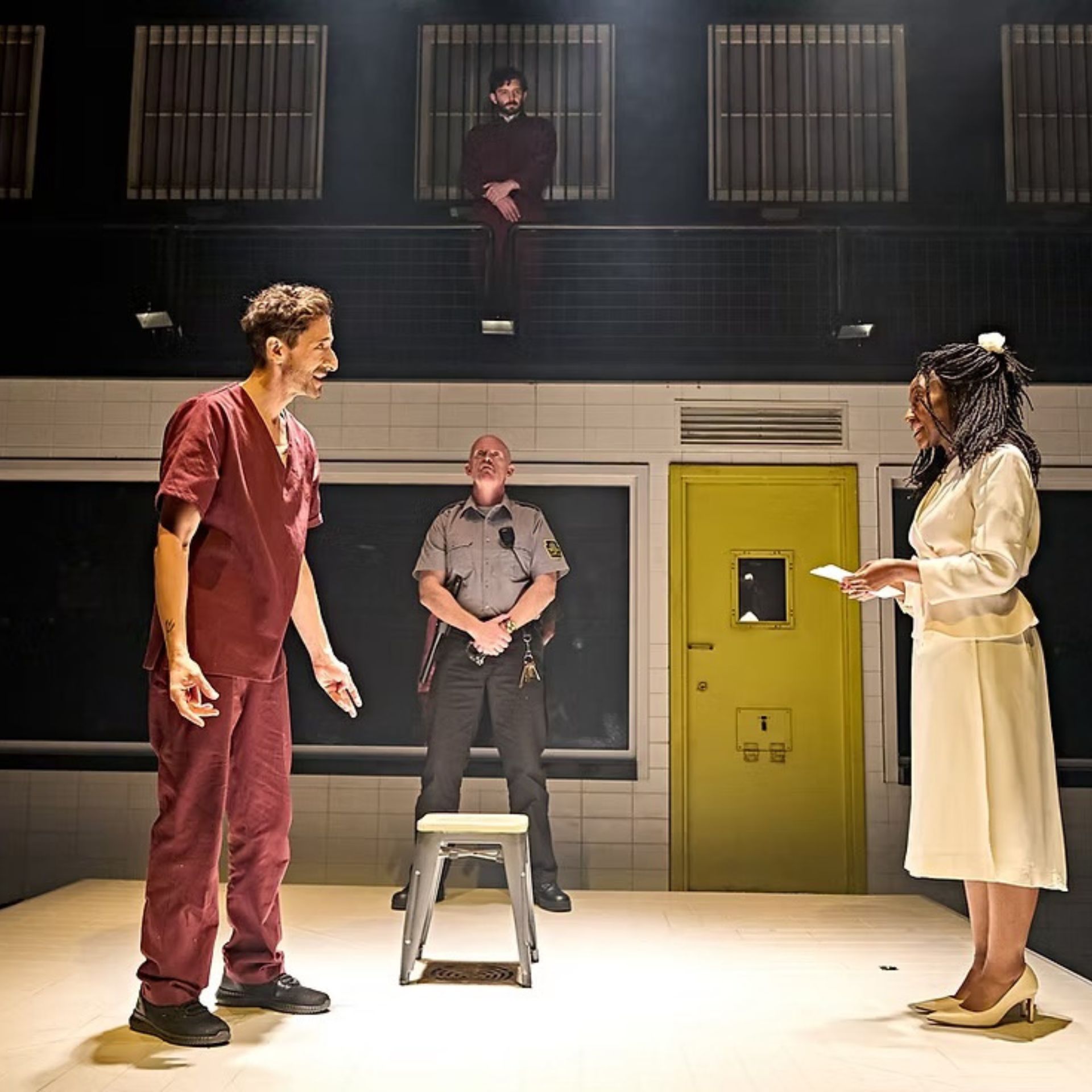 Stage performance with actors in prison-like setting, featuring a guard and dramatic lighting