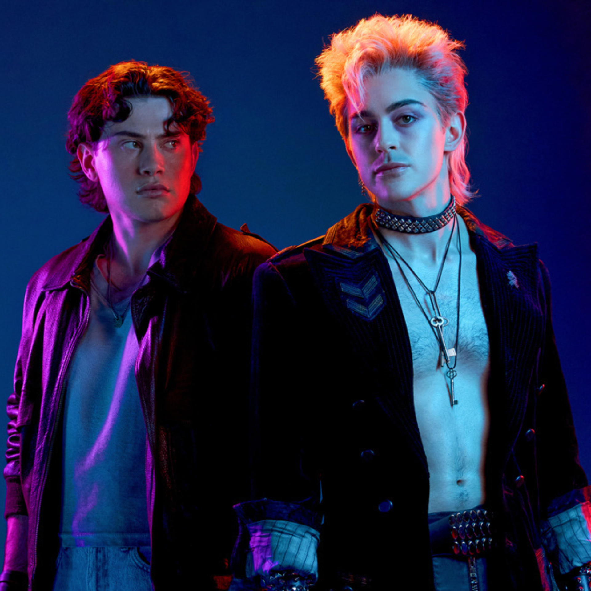Two people stand side by side under pink and blue lighting, one in a leather jacket, the other in punk-inspired outfit with blond mullet