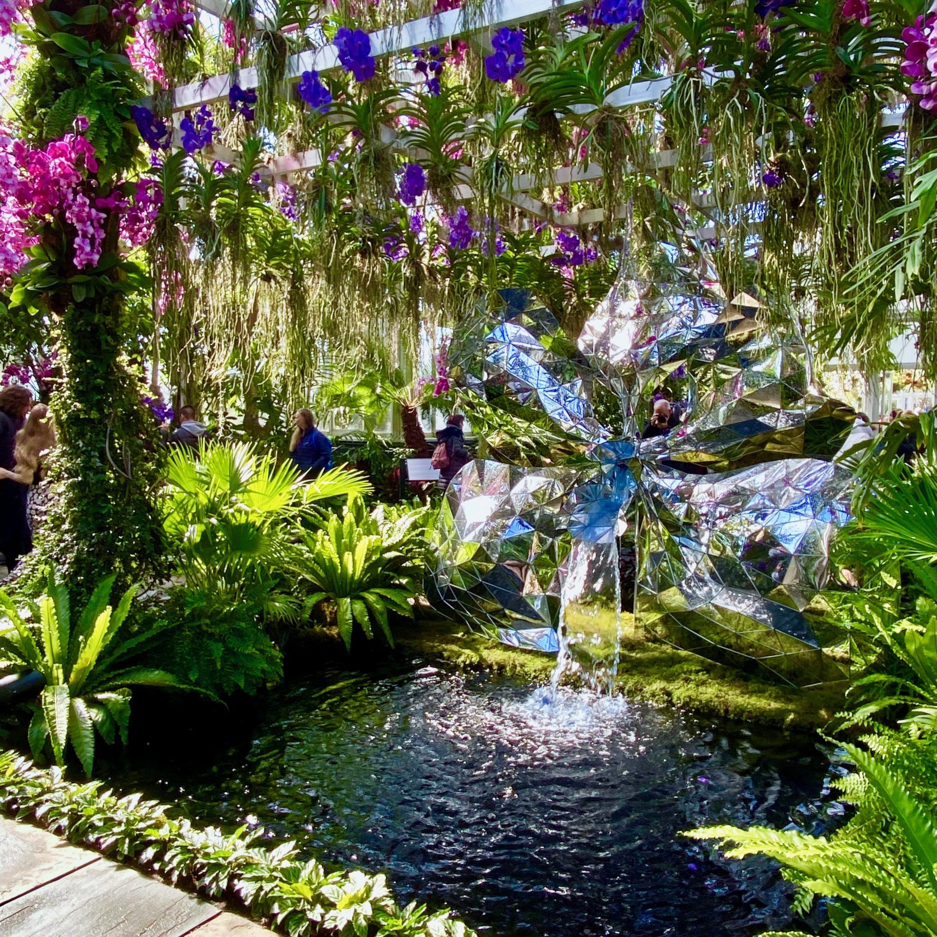 Indoor garden with hanging flowers and a metallic flower sculpture above a pond