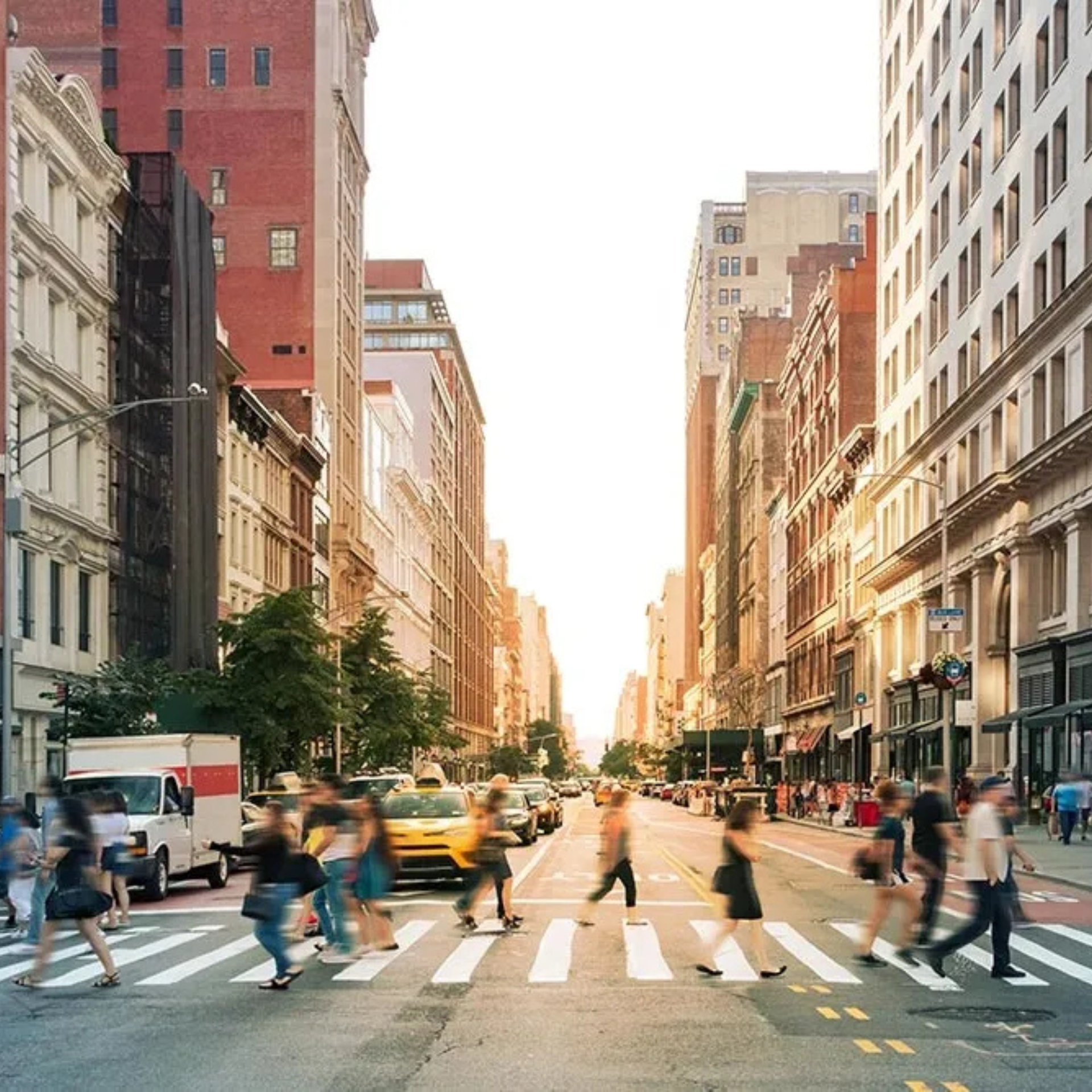 Crowds of pedestrians cross a wide city street, with tall historic buildings and traffic including a yellow taxi
