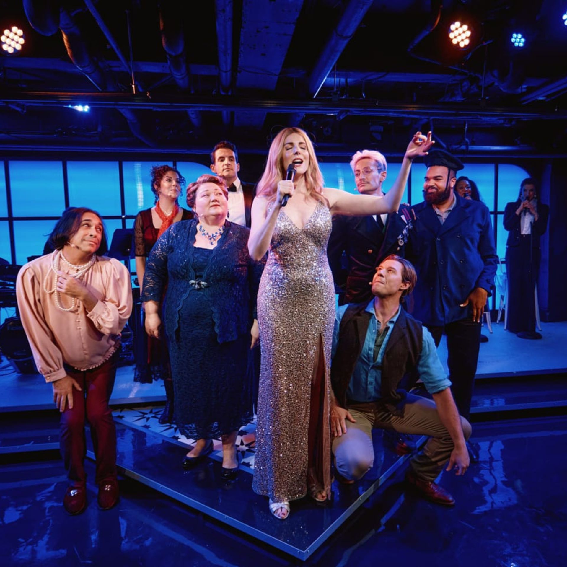 Performer in silver gown sings on stage, surrounded by cast under blue lighting