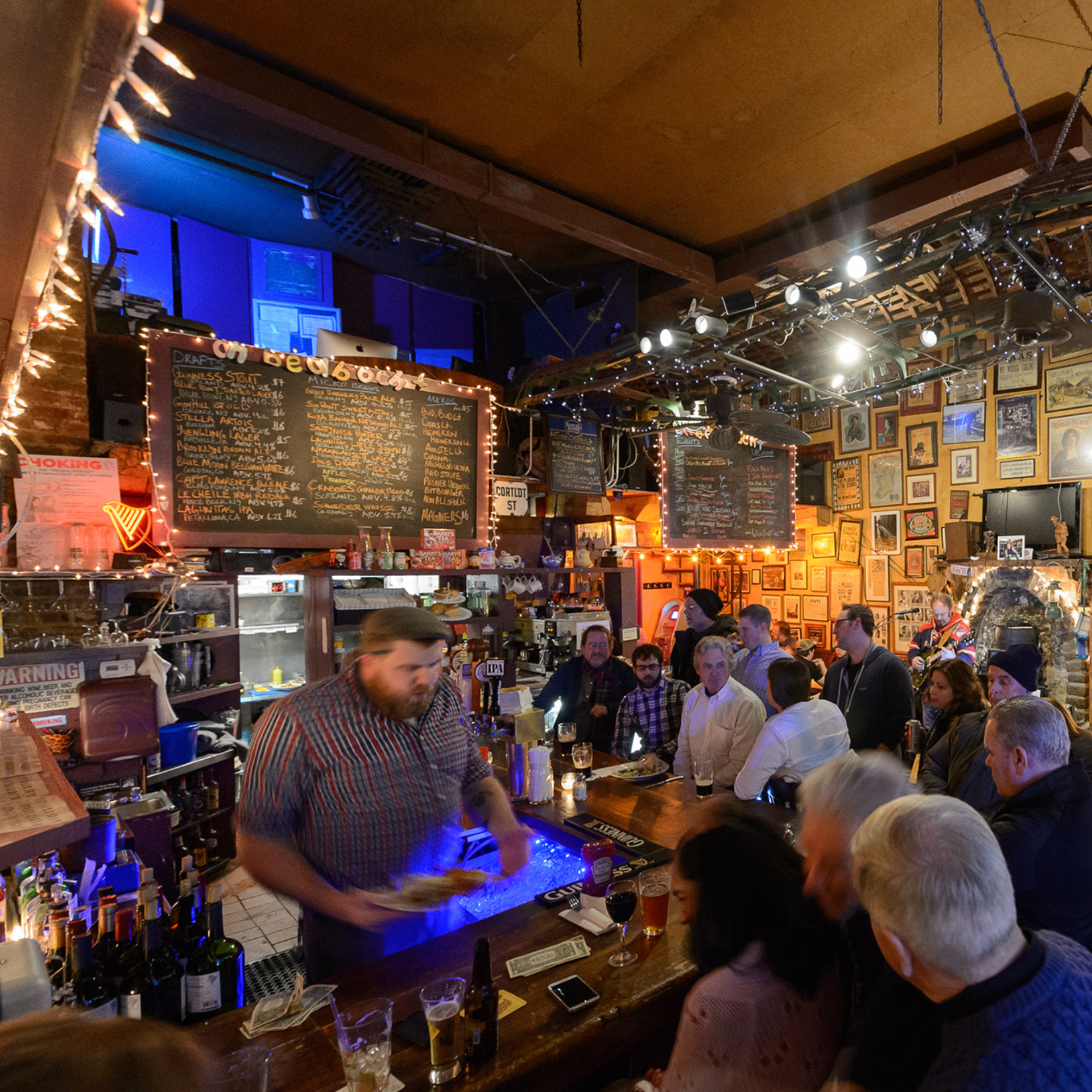 International Jazz Day NYC: Lively bar scene with bartender serving drinks, patrons at wooden counter, and walls covered in framed decor