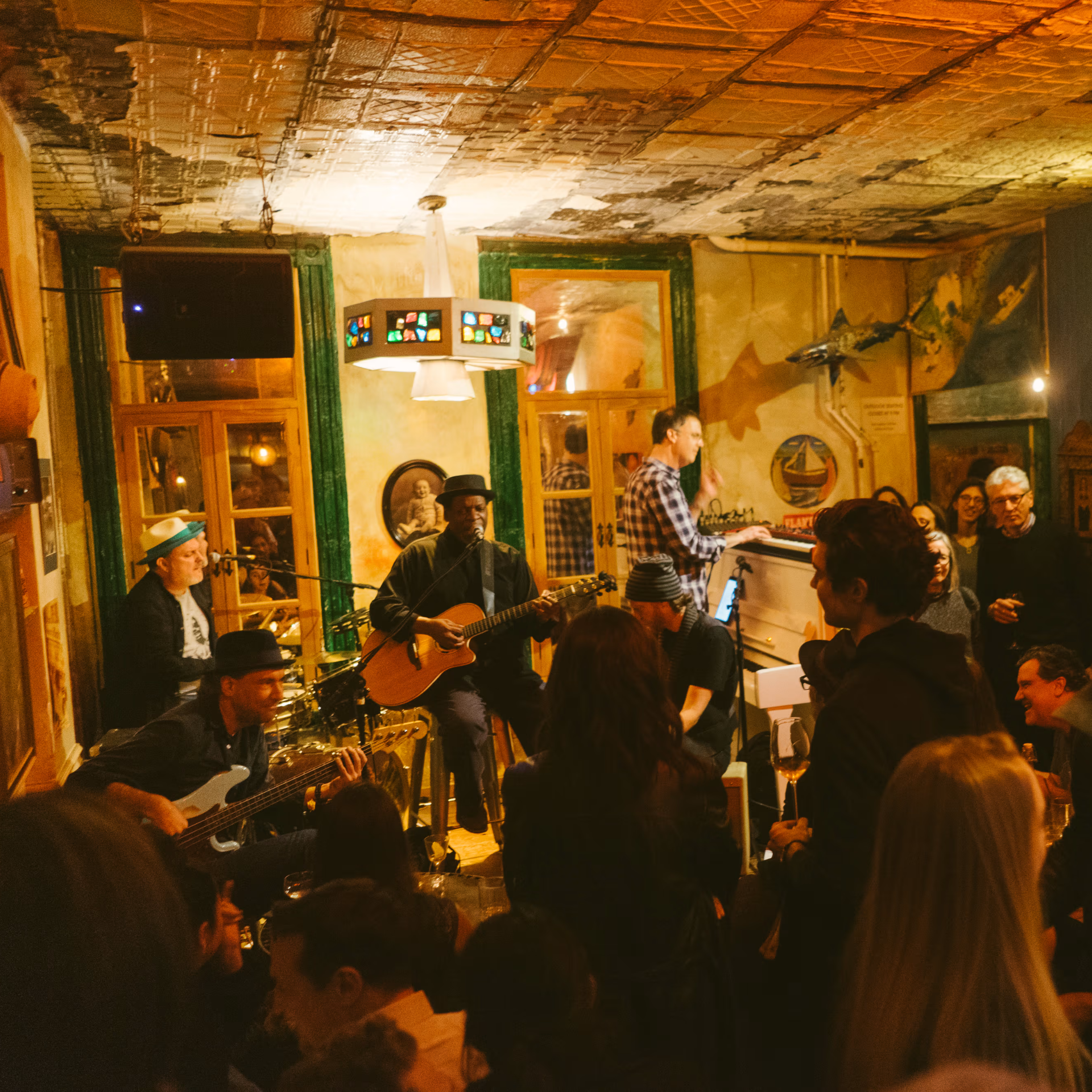 Small bar jam session with guitar, bass, drums, and keyboard, surrounded by audience in warm rustic setting