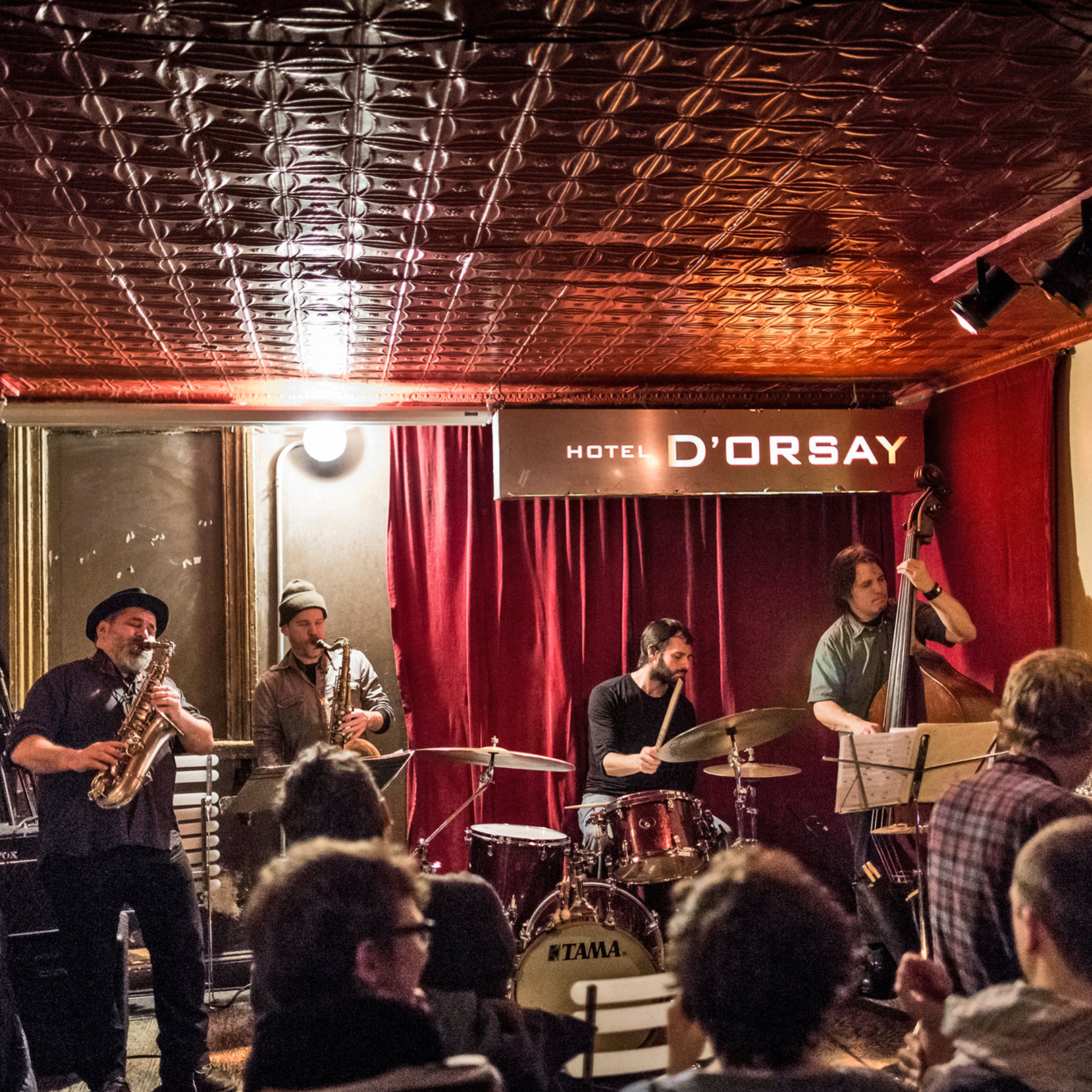 Jazz quartet with two saxophones, bass, and drums performing on stage under red curtains at Hotel D’Orsay