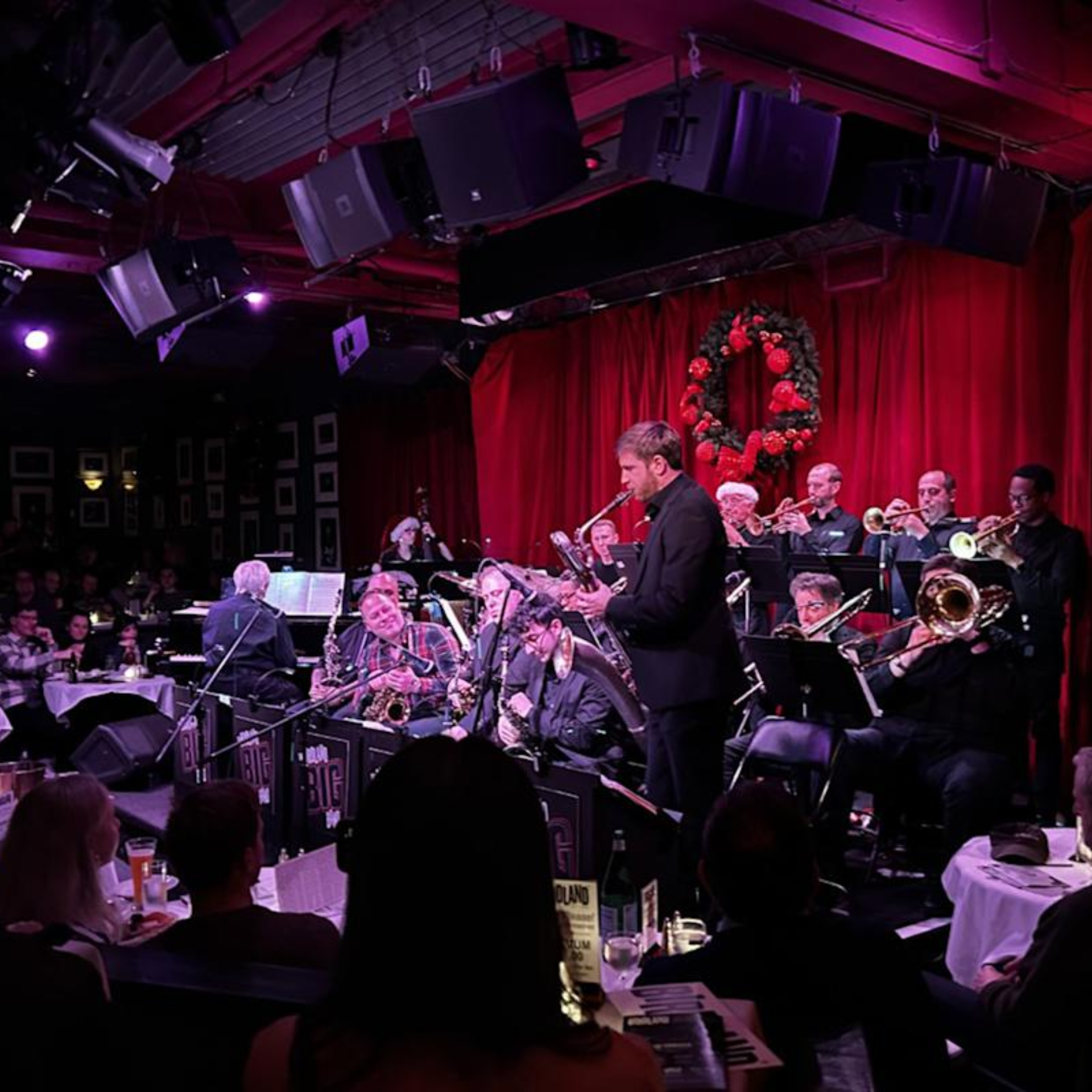 Jazz band with brass and woodwind instruments performing on stage with red curtains and holiday wreath