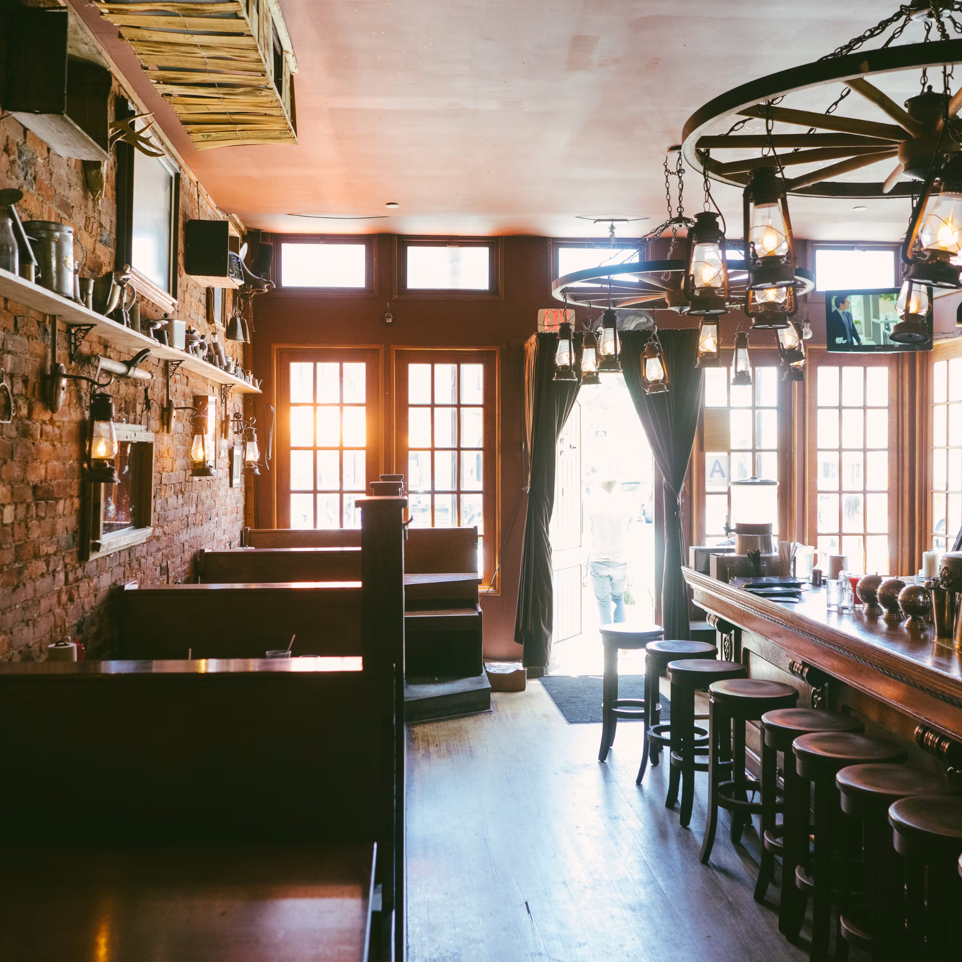 Rustic bar interior with long wooden counter, brick wall with vintage decor, wagon‑wheel chandeliers, and warm sunlight