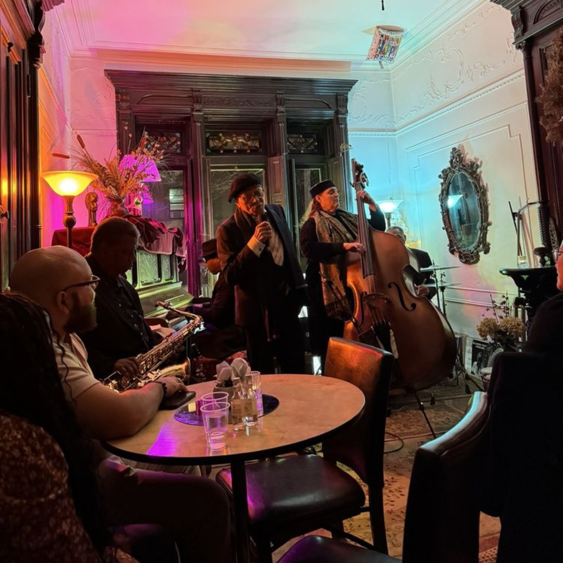 Intimate jazz performance in ornate room with singer, bass, drums, piano, and colorful lighting