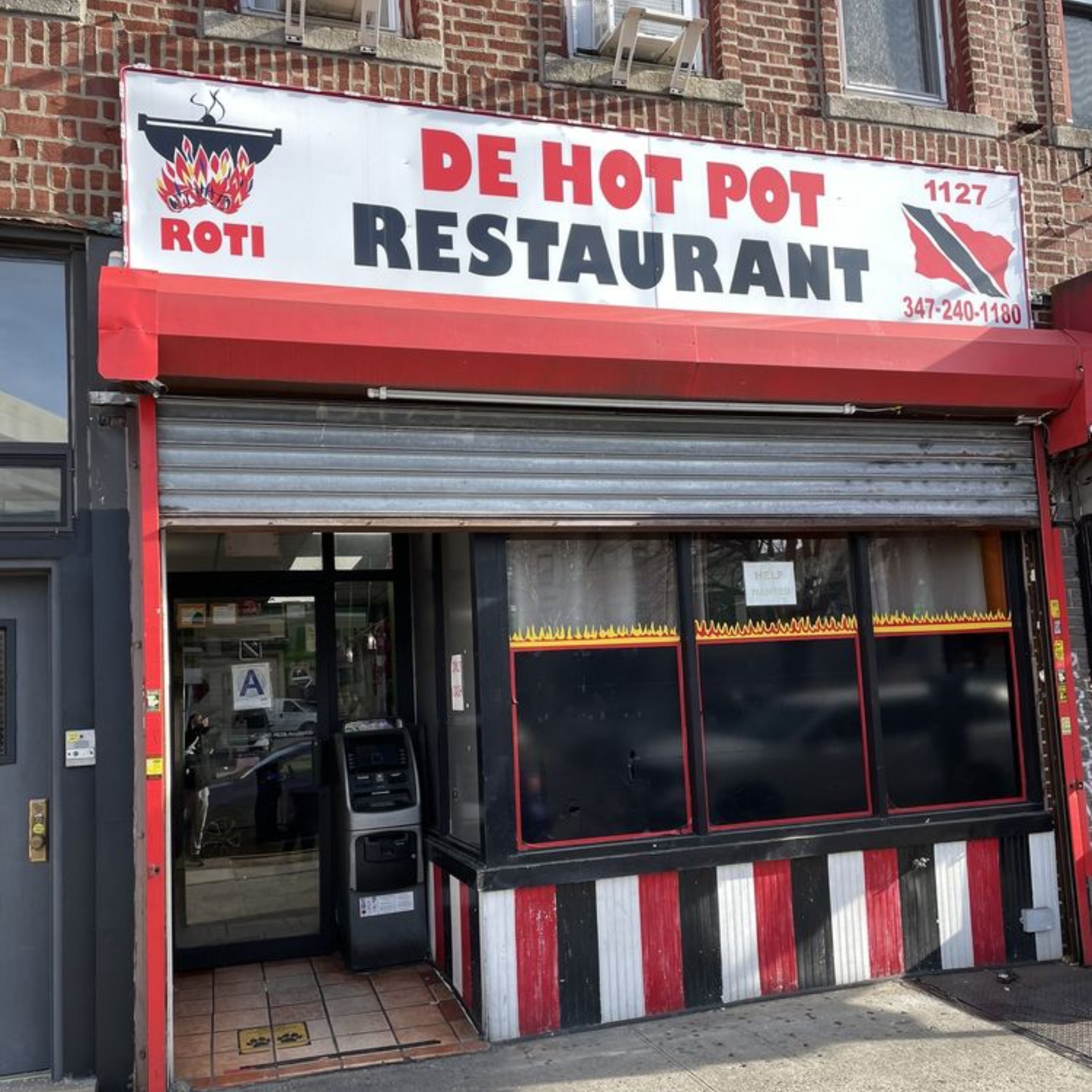 Exterior of Caribbean restaurant “DE HOT POT” with bold sign, Trinidad flag, and partially open entrance