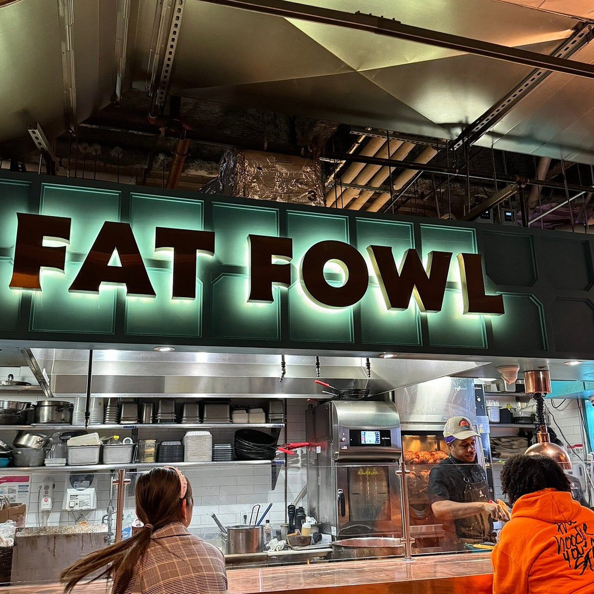 Restaurant counter with illuminated “FAT FOWL” sign, staff working, and customers interacting in a food hall