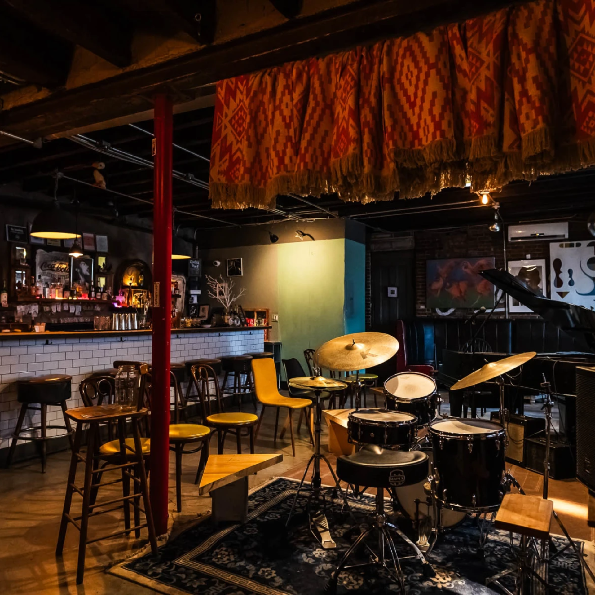 Cozy jazz bar interior with drum set on patterned rug, piano, wooden chairs, and warm lighting