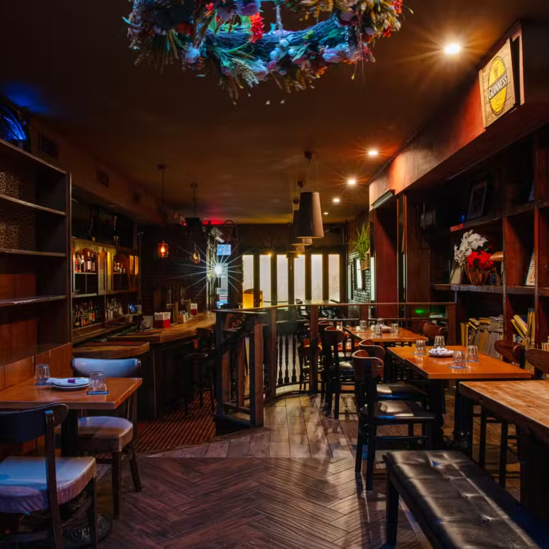 Cozy bar interior with wooden tables, herringbone floor, decorative wreath, and softly lit shelves of bottles
