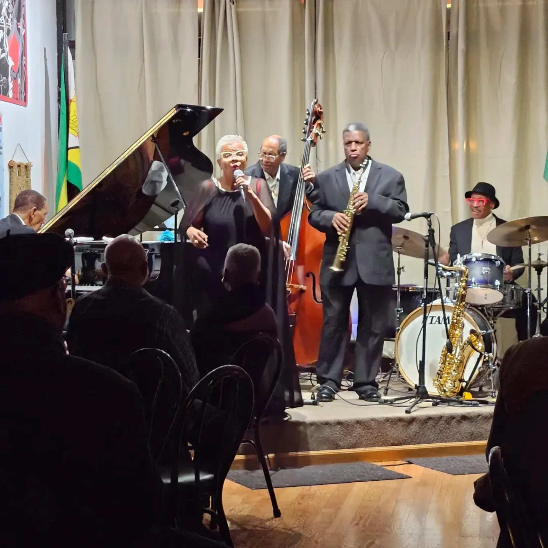 Live jazz performance with vocalist, pianist, bassist, saxophonist, and drummer on stage in warm-lit venue